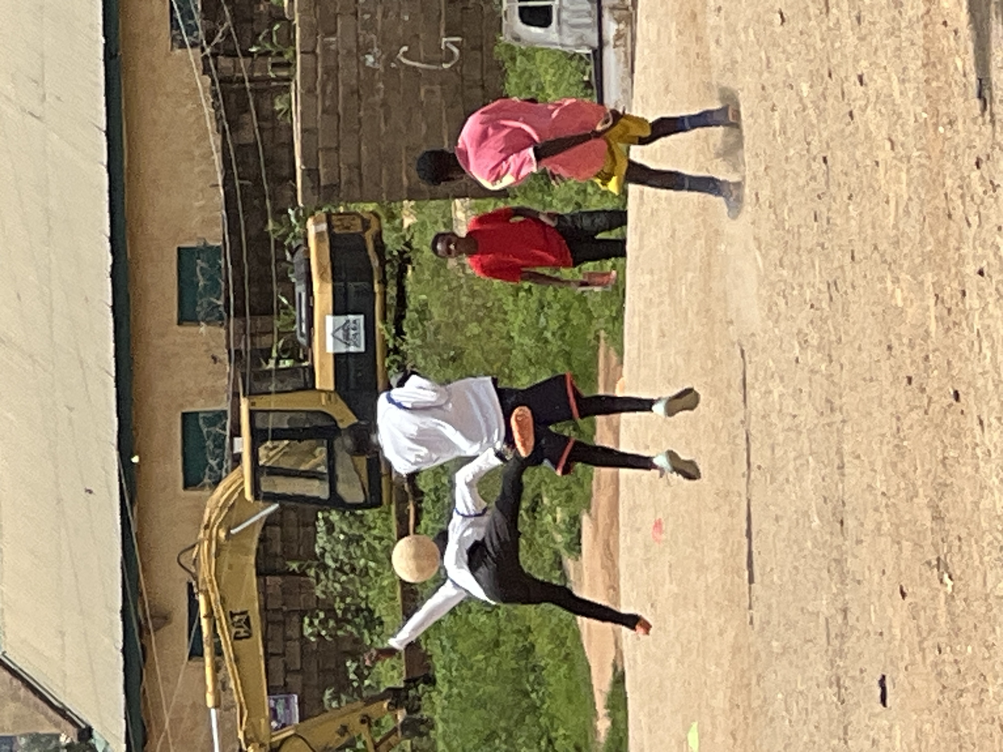 In Jos, Nigeria, three young men are engaged in a soccer game on a dirt field. Two of the individuals are actively playing. One, wearing a white long-sleeved shirt and black pants, is mid-air with his left leg extended, attempting to head a soccer ball. Another player, in a white shirt and dark shorts, stands nearby, also appearing to be in the game. A third individual, in a pink shirt and yellow shorts, is facing away from the action, seemingly observing the game. In the background, a yellow excavator is visible, suggesting ongoing construction or infrastructure work in the area. The environment appears to be a semi-urban setting with buildings and overgrown vegetation surrounding the playing field. The scene captures a moment of athletic activity amidst a backdrop of industrial equipment.