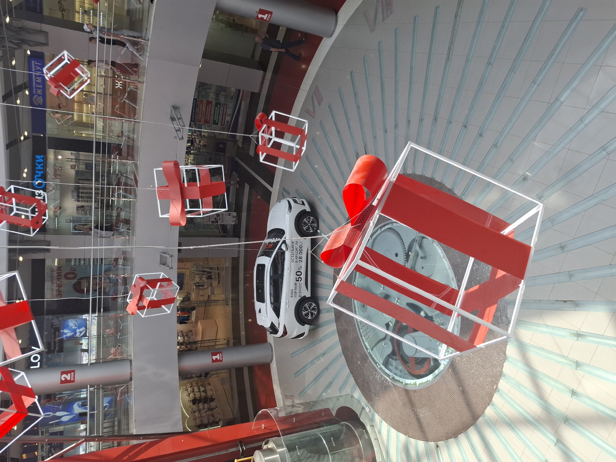 A high-angle indoor shot shows a shopping mall or multi-level commercial space, likely in Izhevsk, Russia, judging by Cyrillic script on signs. Decorations of white wireframe cubes with red ribbons are suspended from the ceiling, spaced throughout the interior. A white car is centrally positioned, likely for promotional purposes. A large, circular structure with visible gears is also present, possibly decorative or functional. Some people can be seen walking on the upper levels. Storefronts with display windows are visible in the background, along with signage for stores like "Zhemchug" jewelry and what appear to be optical services. A red sign with the text "1 этаж" indicates a floor level.