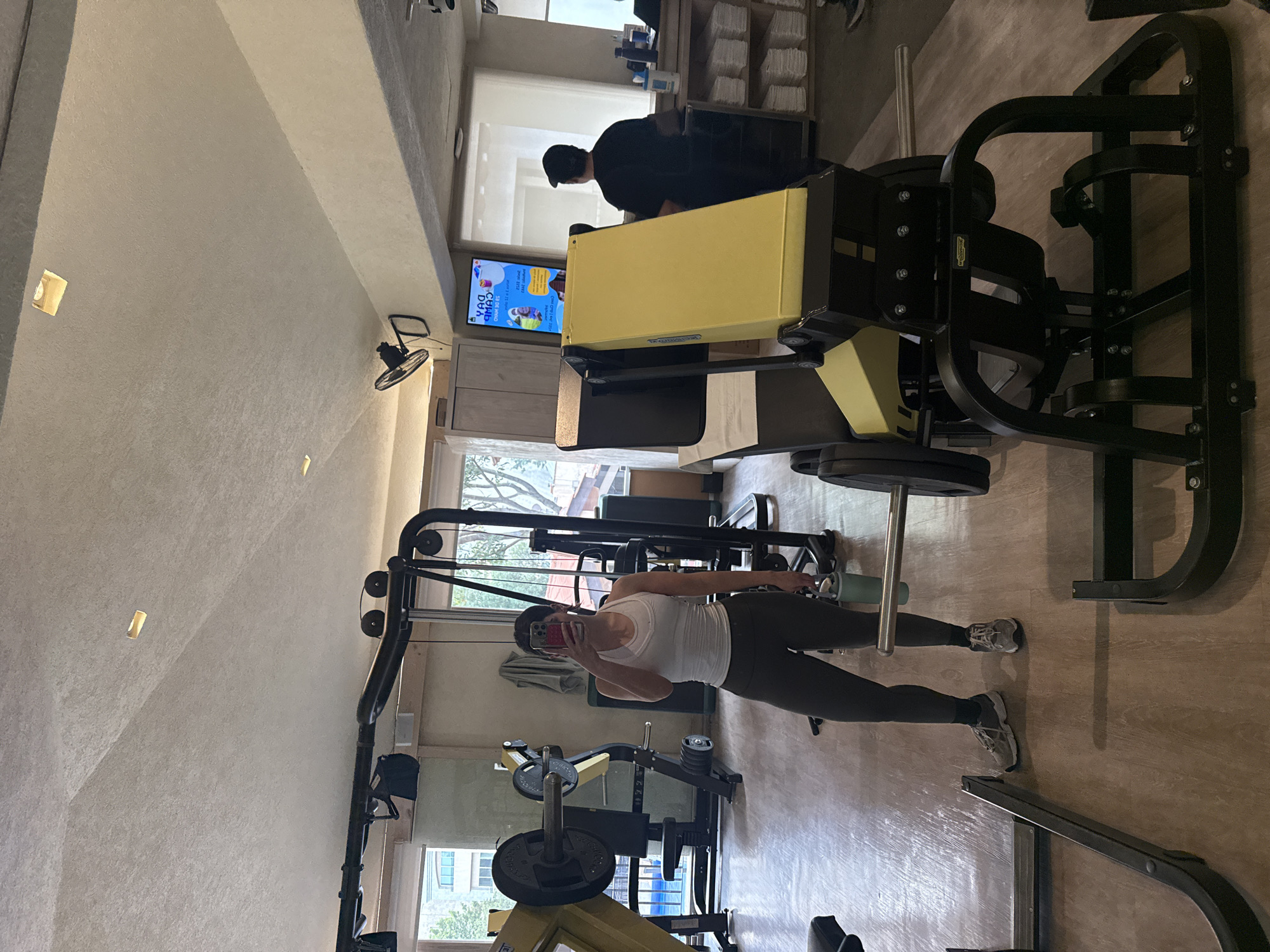 A person is taking a picture in a gym in Huixquilucan de Degollado, Mexico. The person is wearing a white tank top and dark leggings. Gym equipment is visible in the foreground and background. Other people are present in the background. The gym has large windows.