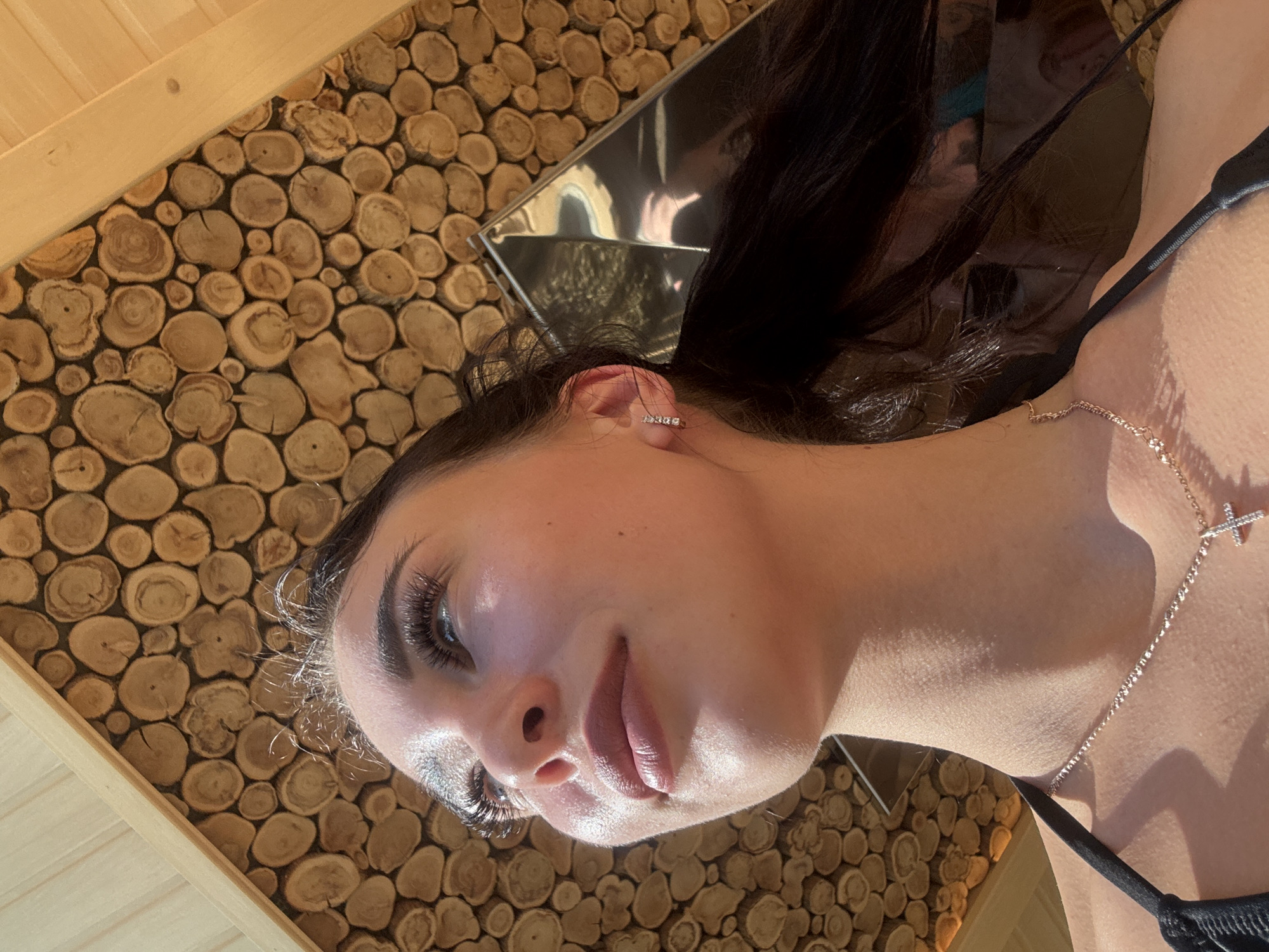 The image is a close-up shot of a woman. She has dark hair, fair skin, and is wearing a necklace with a cross pendant and an earring. She has eyelash extensions and her lips are tinted a light color. She appears to be lying down, as indicated by the surface behind her head and upper back, which is a wall or surface covered in a mosaic-like pattern of small, round wooden pieces. There is also a metallic object behind her head that looks like a frame or a mirror reflecting light. The image does not show enough detail to locate the city or environment in Votkinsk, Russia.