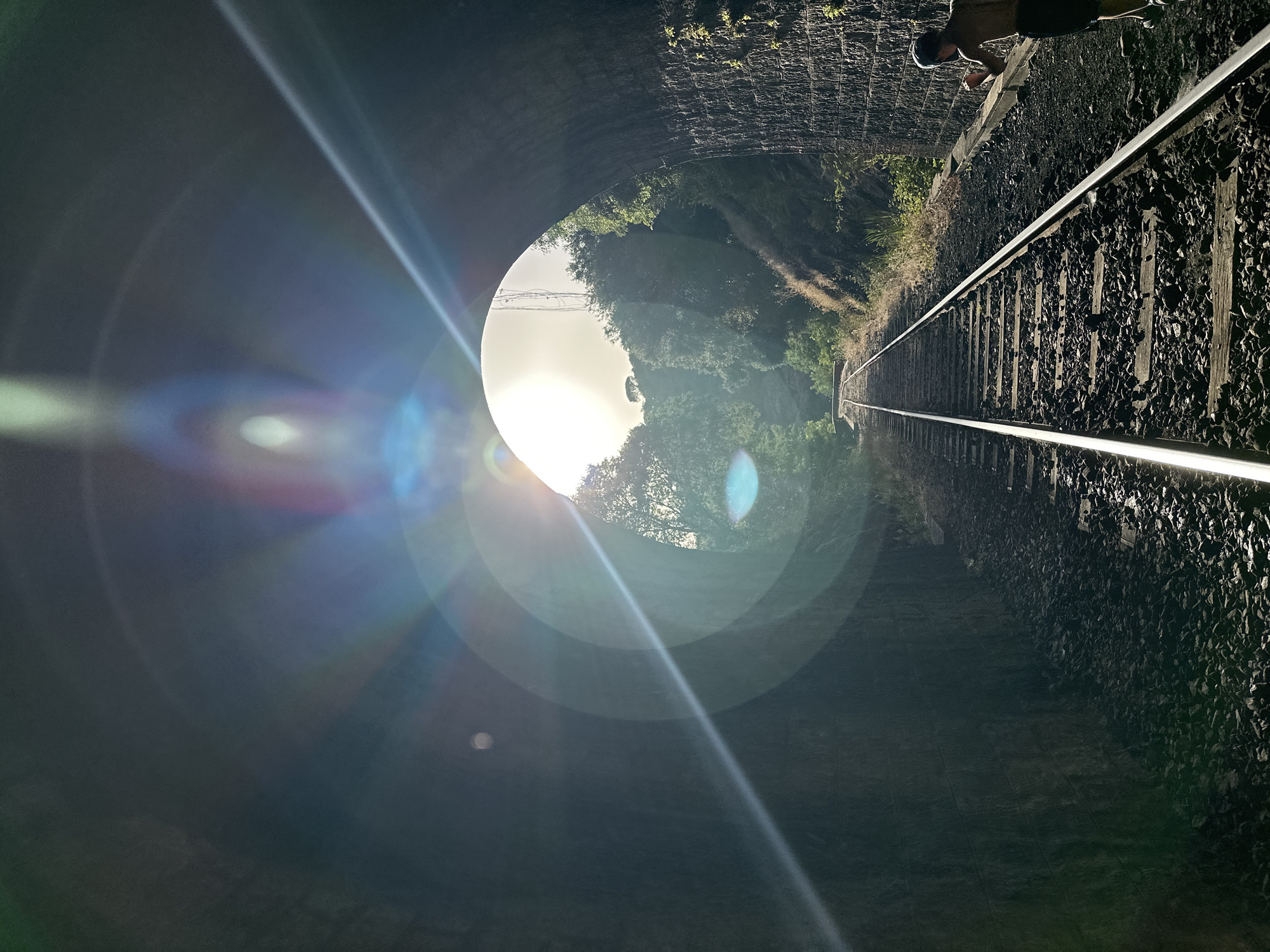 The image shows a view from inside a dark, arched railway tunnel looking outwards towards a bright, sunlit clearing. Railroad tracks lead into the distance, disappearing into the light. The tunnel walls are constructed of rough stone. Trees and vegetation are visible on the slopes outside the tunnel. Lens flare from the sun creates prominent light artifacts, including concentric circles and streaks, across the image. A person, shirtless and wearing dark shorts and a cap, is visible on the right side of the frame, appearing to be running or jogging alongside the tracks. The person's body is mostly in shadow. The scene appears to be in an outdoor, rural environment, with no clear urban indicators.