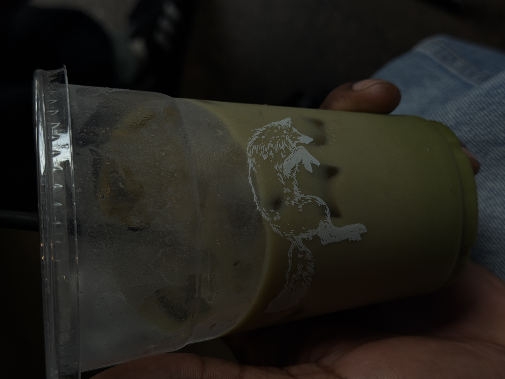 A person's hand is holding a clear plastic cup filled with a light green beverage and ice. A white graphic of a wolf is visible on the side of the cup. The background is dark and out of focus, with a hint of denim fabric visible at the bottom. The setting is indoors, likely a cafe or restaurant. The image does not contain information to determine the location in Columbus, Ohio, USA.