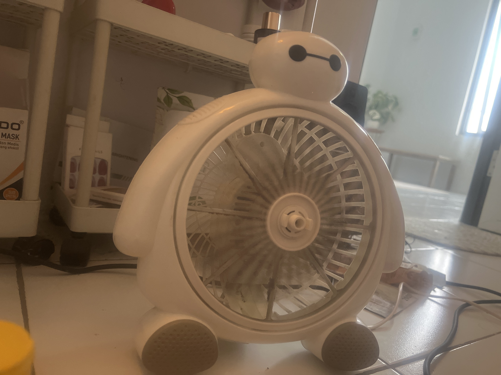 A white, Baymax-shaped fan is positioned on a tiled floor in a dimly lit room. The fan's head features two black circles for eyes and a black horizontal line for a mouth, mimicking the character's face. Its body is circular, with the fan blades visible behind a white grate. The fan has two small, textured feet. Behind the fan, a white shelf unit with multiple shelves is visible, holding various items including boxes and bottles. To the right of the fan, a large window allows natural light to enter the room, illuminating a plant in the background. A black power cord runs from the fan towards the right side of the frame. The floor is tiled with a light-colored, possibly beige or cream, pattern.