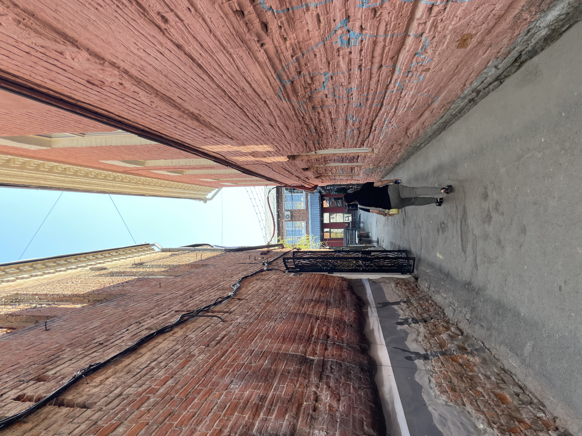 A person walks away from the viewer in a narrow alleyway between brick buildings. The alley is paved with concrete. The buildings are brick, with the left building appearing older and more weathered. The building on the right is red brick. The perspective is looking up. A wrought iron balcony railing is attached to the bottom left building's wall. The sky is visible at the top of the alleyway. Based on the location cues from the prompt, this alleyway is located in Izhevsk, Russia.