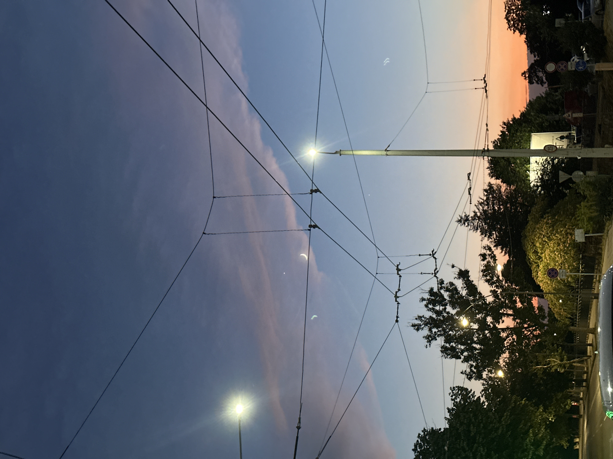 The image depicts a twilight scene in the city of Ruse, Bulgaria, as indicated by the presence of trolleybus wires crisscrossing the sky, a common feature of the city's public transportation system. The sky transitions from a deep blue overhead to a soft orange and pink hue near the horizon, suggesting the sun has recently set. Several streetlights are illuminated, casting a bright glow. In the sky, a crescent moon is visible, along with what appear to be two faint celestial bodies or atmospheric phenomena. The foreground shows a street with trees lining the sides, and the tops of vehicles are visible, implying traffic. A few traffic signs are present on the right side of the image. The overall impression is a serene urban evening, with the overhead wires creating a distinct urban pattern against the colorful sky.