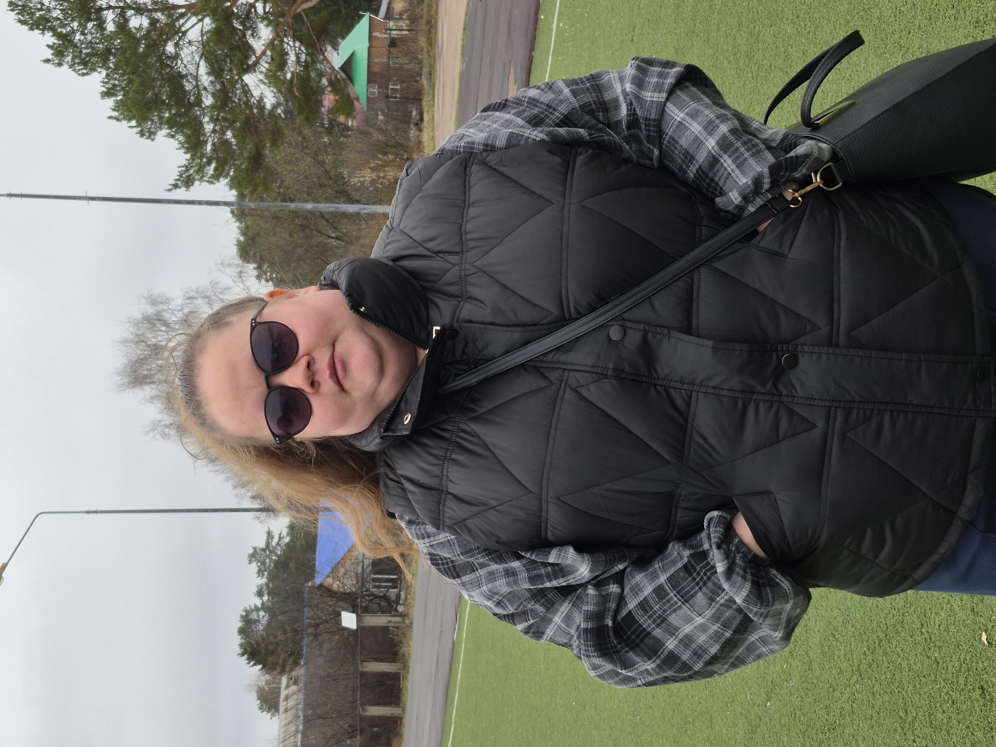 A medium shot captures a woman standing outdoors in what appears to be a sports field in Chaikovsky, Russia. She is looking directly at the camera with a slight smile and is wearing dark sunglasses. Her attire consists of a black quilted vest, a gray and black plaid long-sleeved shirt, and dark trousers or jeans. A black shoulder bag with a gold clasp is visible hanging from her right shoulder. Her light-colored hair frames her face. Her left hand is casually placed in the pocket of her vest.
The background shows a large expanse of artificial green turf, possibly a football or track field, with a narrow asphalt path running alongside it. Beyond the field, there are bare deciduous trees and evergreen pine trees under an overcast sky, suggesting it might be late autumn or early spring. Several buildings are visible in the distance, including one with a distinctive green-sloped roof and another with a blue tarp covering part of it. A tall light pole extends upwards on the left side of the frame. The overall atmosphere is cool and muted due to the cloudy weather. No visible text is present in the image.