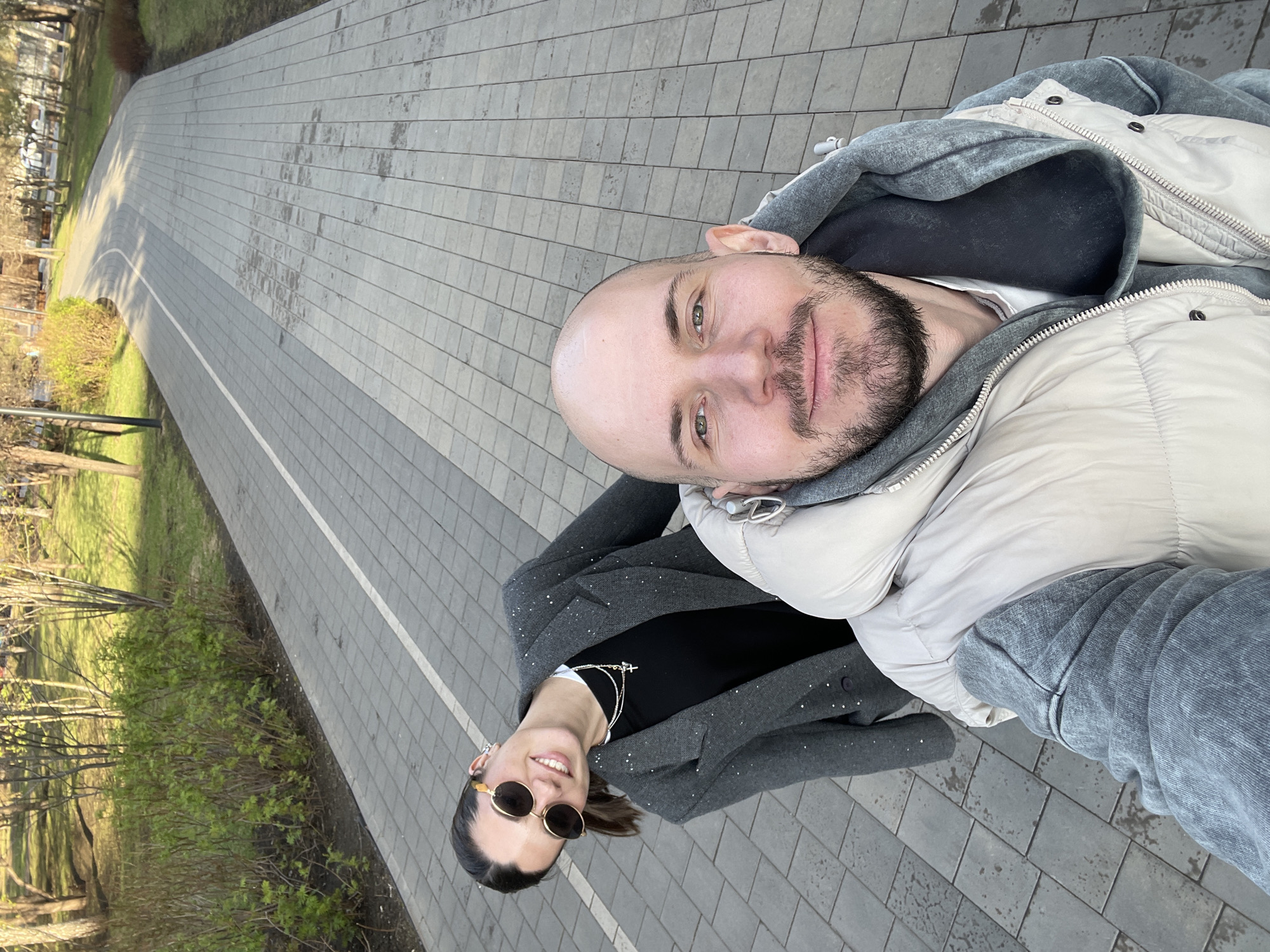 A selfie photograph shows a man and woman posing on a gray brick pathway. The man is fair-skinned with a shaved head and a beard. He wears a gray hooded sweatshirt layered under a light beige puffer jacket. The woman, also fair-skinned, stands slightly behind and to the left of the man. She is wearing sunglasses, a black top, and a gray blazer with sparkly embellishments. Two necklaces, one with a cross pendant, are visible. The pathway is lined with green foliage and trees to the left, and open grass on the right. The path curves into the distance. The photo appears to be taken in the city of Krasnoyarsk, Russia.