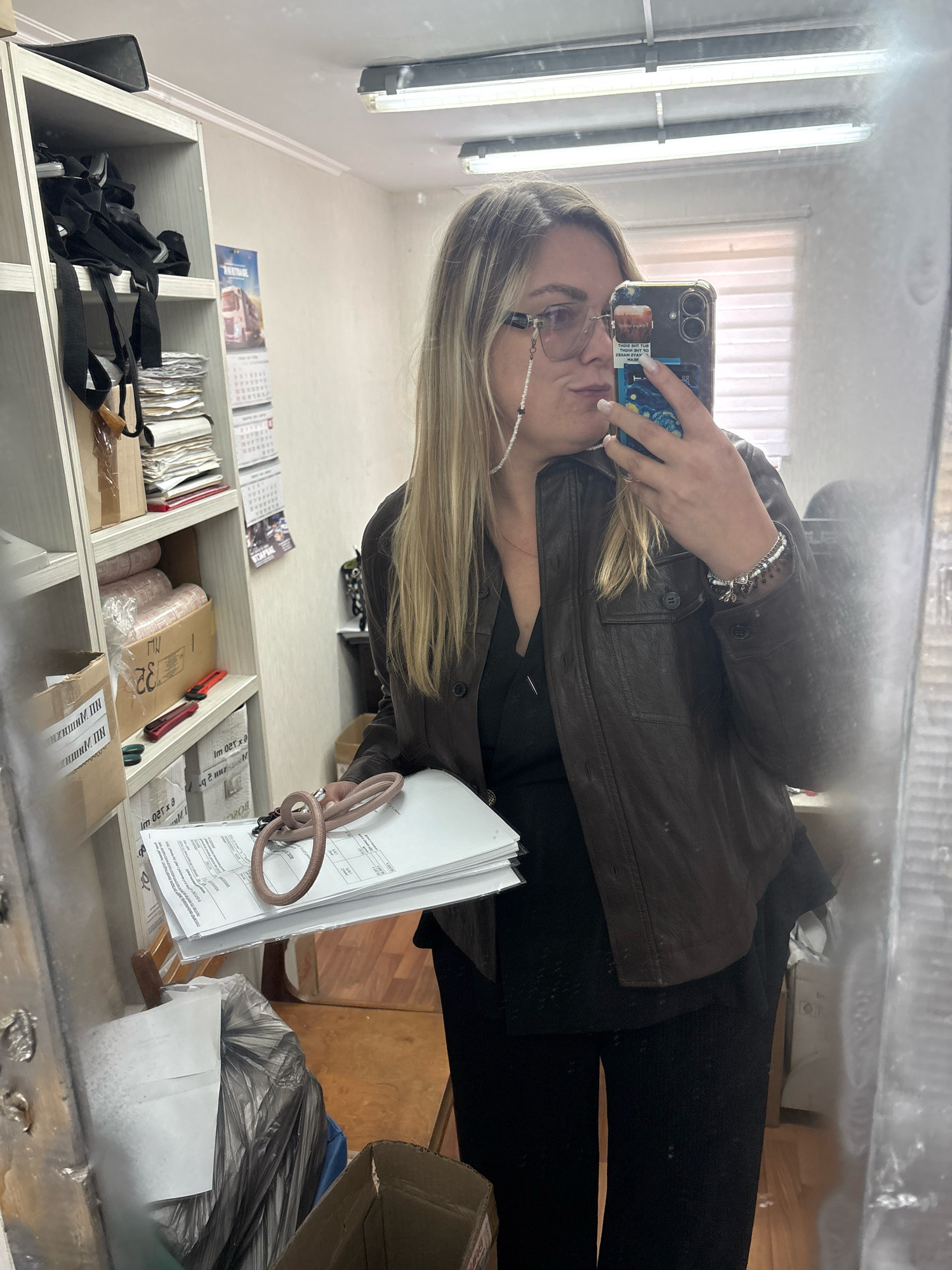 A woman with blonde hair is taking a mirror selfie in what appears to be an office or storage room. She is wearing glasses with a beaded chain, a brown leather jacket, and black clothing. In her right hand, she holds a smartphone, the screen of which is visible. In her left hand, she holds a stack of papers with a pink cord wrapped around them. Behind her, shelves are filled with various items, including boxes, papers, and black straps. A calendar with images of trucks is attached to the wall. The room has fluorescent lighting and the floor appears to be wood. The reflection in the mirror suggests the location is not in the city of Noviy, Russia, but rather an indoor environment that could be anywhere.