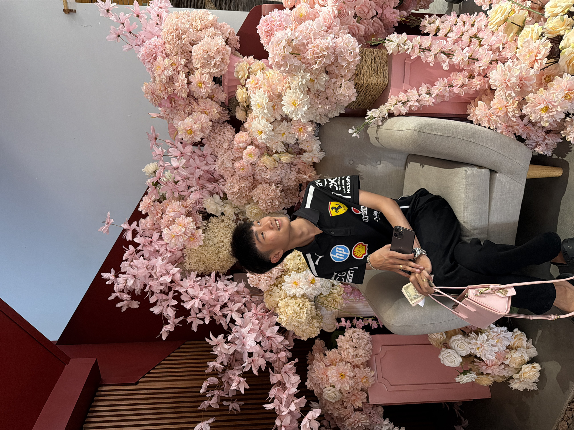 A man in a black shirt and pants reclines on a grey armchair, surrounded by pink and white artificial flowers. He is holding a phone in one hand and a small pink purse with cash protruding in the other. The backdrop includes a wall painted in maroon and greyish-blue, and wooden slats. The photo appears to be taken in Quy Nhon Ward, Vietnam.