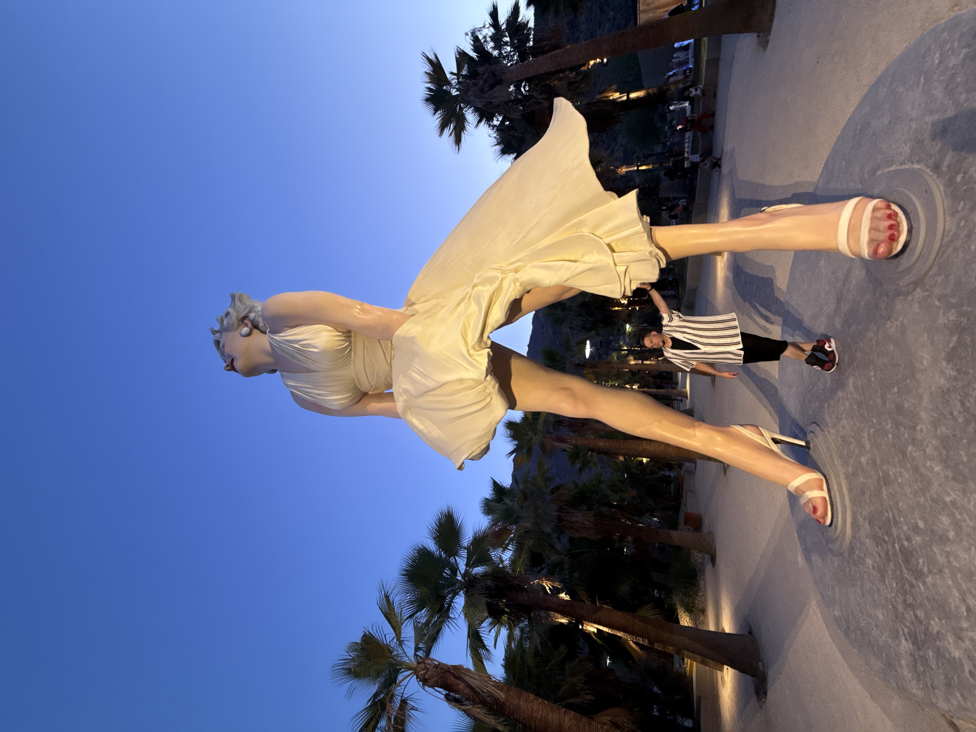In Palm Springs, California, a large sculpture of Marilyn Monroe stands in an outdoor plaza. The statue is a rendition of her iconic pose from the movie "The Seven Year Itch," with her white dress blowing up around her legs. A person stands in front of the statue, looking up at it. The scene takes place at dusk, with a clear blue sky and palm trees in the background. The ground is paved, and there are lights illuminating the area.