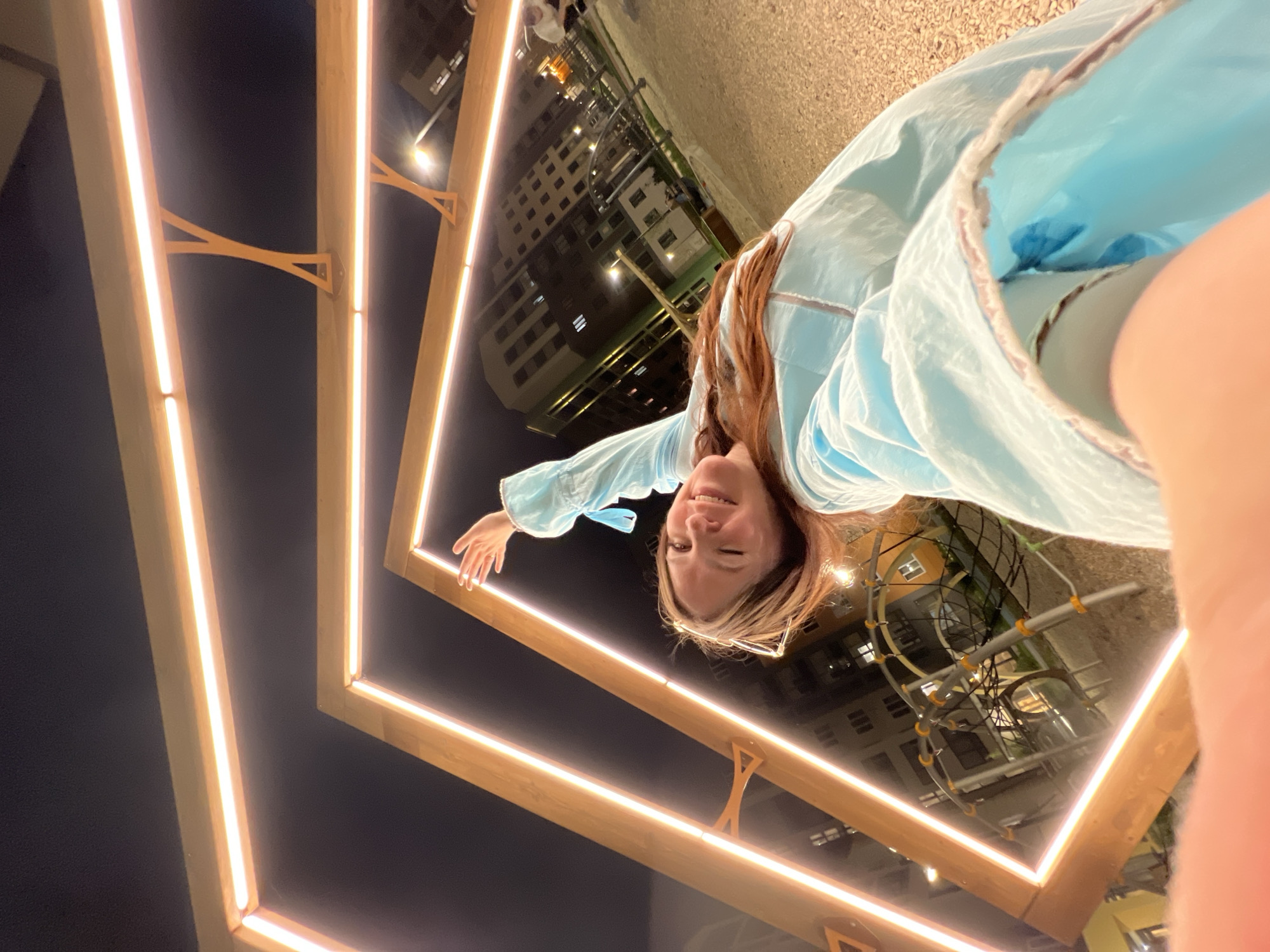 A woman with long reddish-brown hair is posing in front of illuminated square frames, likely an art installation or decorative lighting structure. The background suggests an urban environment at dusk or night, with building lights visible. The location is implied to be Pervomaysky, Russia. The woman is wearing glasses and a light blue garment, possibly a shirt or jacket. Her expression is cheerful, and she is extending her hand towards the frame.