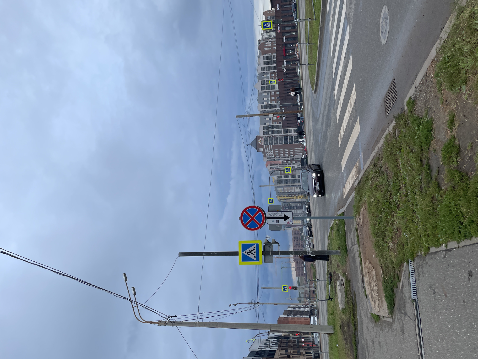 This image depicts a cloudy day in the city of Izhevsk, Russia, likely in a newly developed urban area with modern multi-story apartment buildings and commercial establishments. The foreground shows a wet asphalt road with a clearly marked pedestrian crossing. To the left of the road, there is a concrete sidewalk with patchy green grass. A tall utility pole with streetlights and electrical wires is prominent on the left side of the image.
Several traffic signs are visible, including a pedestrian crossing sign, a "no stopping" sign with a red circle and a diagonal cross, and a directional arrow sign. A dark-colored sedan is driving across the pedestrian crossing. In the middle ground, there are more traffic lights and signs, as well as several parked cars and some pedestrians. The background is dominated by the architecture of apartment buildings and commercial spaces, some of which feature prominent signage. Construction cranes are visible in the distance, suggesting ongoing development in the area. The overall atmosphere is overcast and somewhat muted, consistent with a cloudy day.