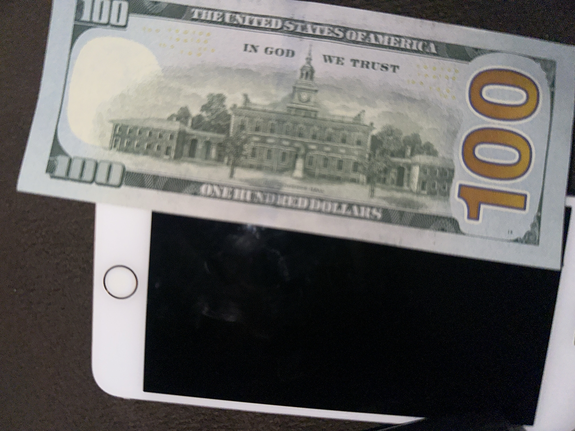 A U.S. one hundred dollar bill is placed on top of a white smartphone. The background is a dark, textured surface.