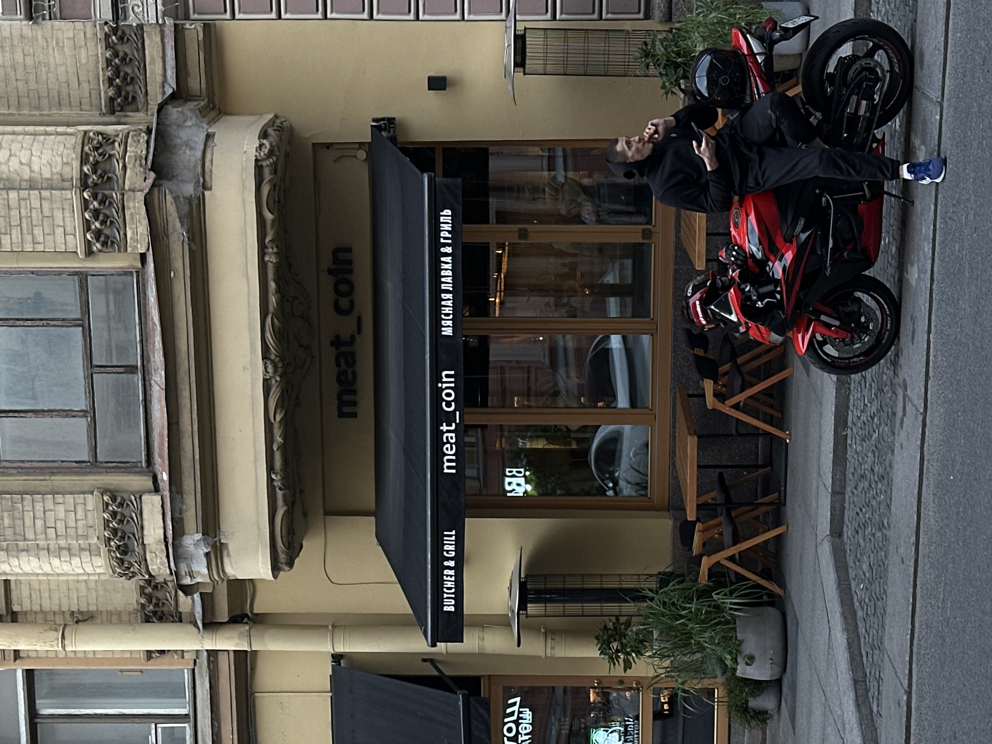 The image shows a man sitting on a red and black motorcycle parked on a sidewalk in front of a building in Saint Petersburg, Russia. The man is wearing a black hoodie and black pants, with headphones on and is holding a phone in his left hand and a piece of food in his right hand, which he appears to be eating. The motorcycle is a sportbike with a black helmet resting on the rear seat.
The building has a yellow facade with architectural details and large windows. Above the entrance, there is a black awning with the text "meat_coin" and "BUTCHER & GRILL" in English, and "МЯСНАЯ ЛАВКА & ГРИЛЬ" in Russian, indicating a butcher shop and grill. Outside the establishment, there are outdoor seating arrangements with wooden tables and chairs. Planters with greenery are placed near the entrance and along the sidewalk. The sidewalk is paved with cobblestones and asphalt, and a street is visible in the foreground. The overall scene suggests a casual urban setting.