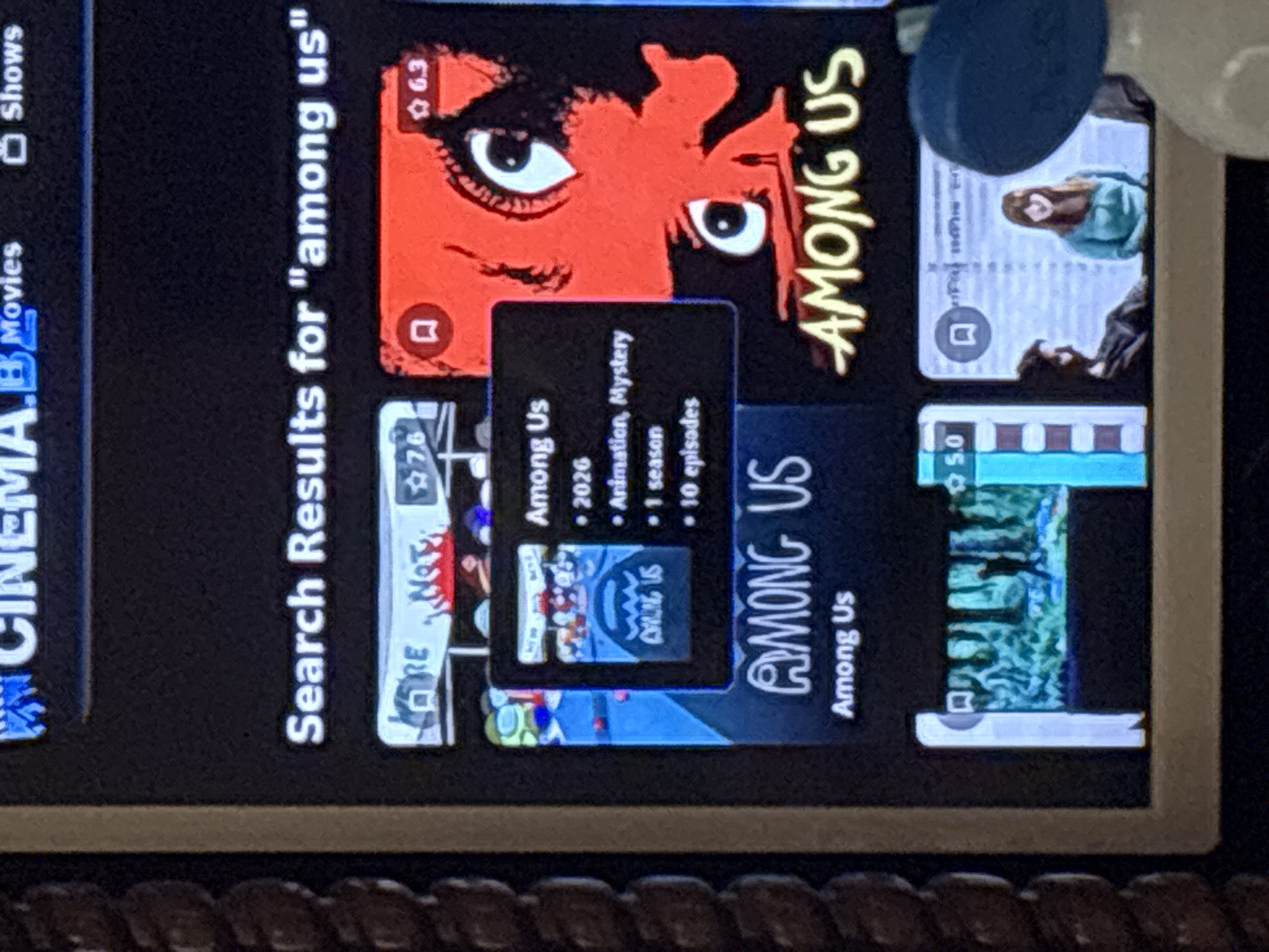 The photograph captures a digital screen displaying search results for "among us" on what appears to be a streaming or media application interface. This scene is observed in Burgas, Bulgaria.
The screen's upper section features a partial header, "CINEMA.B Movies Shows," indicating a media platform. Below this, the prominent text "Search Results for 'among us'" is displayed.
The central part of the screen presents multiple media entries in a grid layout. A featured result, foregrounded with an informational overlay, shows a poster for "Among Us" with various colorful characters against a blue background, dated "2026," categorized as "Animation, Mystery," and detailing "1 season" with "10 episodes." To its right, another prominent result features a red poster with two large, stylized white eyes and a small dark silhouette, titled "AMONG US" in yellow text, bearing a rating of "6.3." A partially visible entry to the far left has a rating of "7.6" and fragments of text "RE Not."
In the lower portion of the screen, two additional search results are partially visible. The leftmost shows a dark, forested scene with a human silhouette, rated "5.0." The rightmost depicts a man in shadow and a woman in a light blue top.
The screen is framed by a light-colored bezel. To the left of the screen, a dark, braided or woven object is visible, likely part of furniture. A blurry, light-blue object, possibly a person's shoulder or clothing, is present in the lower right foreground, suggesting proximity to the device. The screen itself is illuminated, providing the primary light source within the immediate photographic frame.