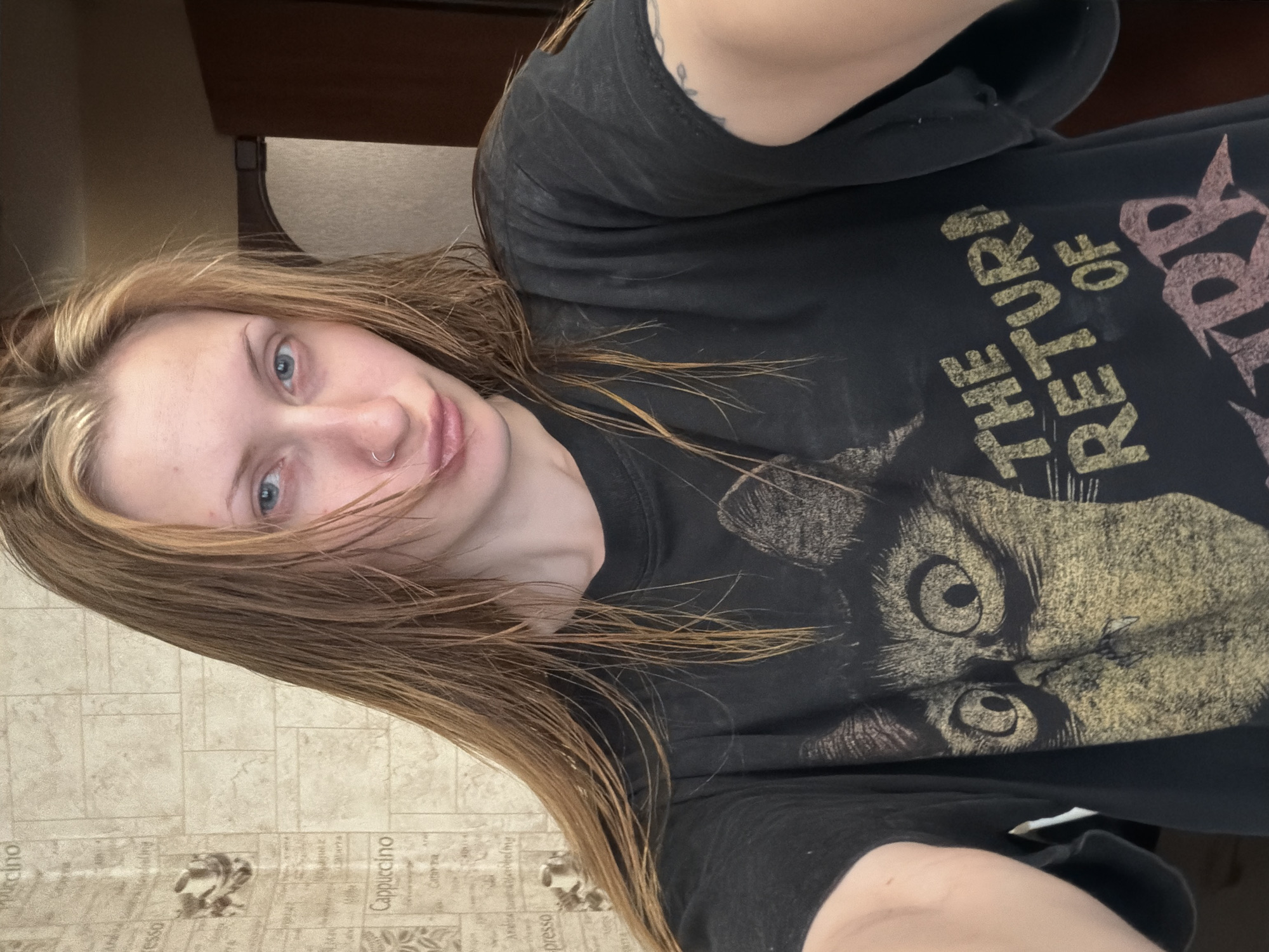 The image is a selfie taken by a young woman with long, wavy blonde hair. She is wearing a black t-shirt with a graphic print of a cat's face and the text "THE RETURN OF". The woman has blue eyes and a nose ring. She is looking directly at the camera with a neutral expression. Her hair is slightly damp, suggesting she may have recently showered or been out in the rain.
The background of the image is out of focus, but appears to be an indoor setting. There is a patterned wallpaper visible on the left side of the image, with a repeating design that includes text that appears to be coffee-related, such as "Cappuccino" and "Espresso". A piece of furniture, possibly a dresser or shelf, is visible in the upper portion of the image.
The lighting in the image is soft and diffused, indicating it might be daytime. The overall mood of the image is casual and relaxed. The woman is positioned in the center of the frame, with her face and upper body prominently displayed. The image is oriented in a portrait aspect ratio. The visible text on the t-shirt, "THE RETURN OF", is in a distressed, vintage font.