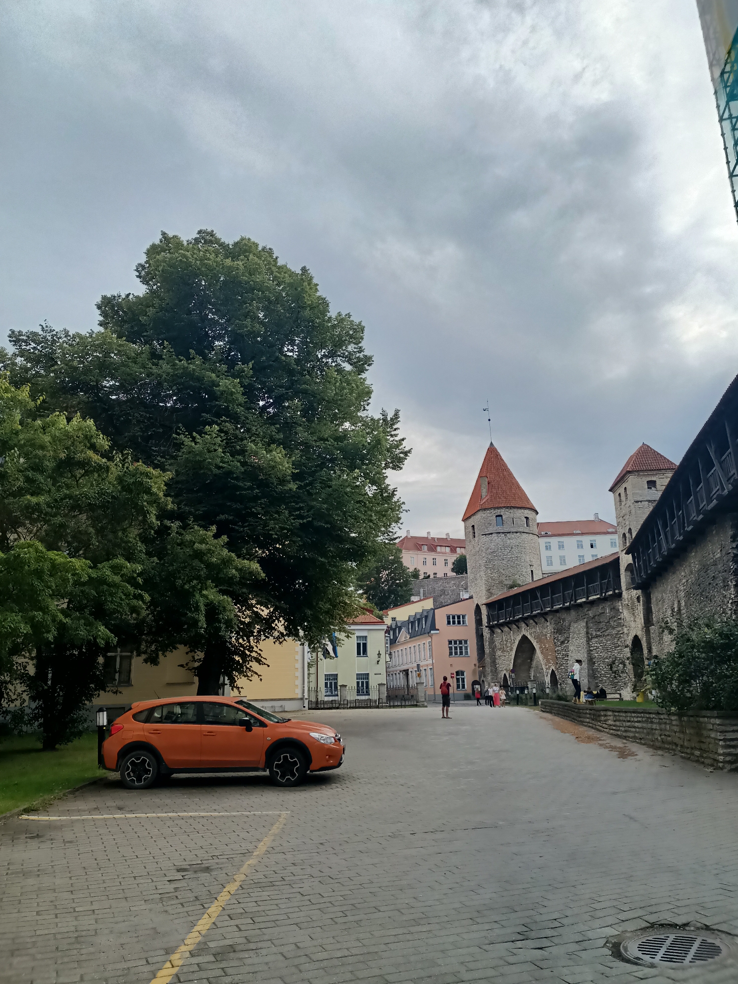 In Tallinn, Estonia, the image depicts a street scene with historical architecture. An orange Subaru XV is parked in a cobblestone lot in the foreground. To the left, a large, leafy tree provides shade. Beyond the lot, the street opens up to reveal a section of Tallinn's medieval city wall, complete with towers and wooden walkways. Several people are visible walking along the street and near the wall. Buildings with pitched roofs line the street in the background. The sky is overcast with grey clouds.