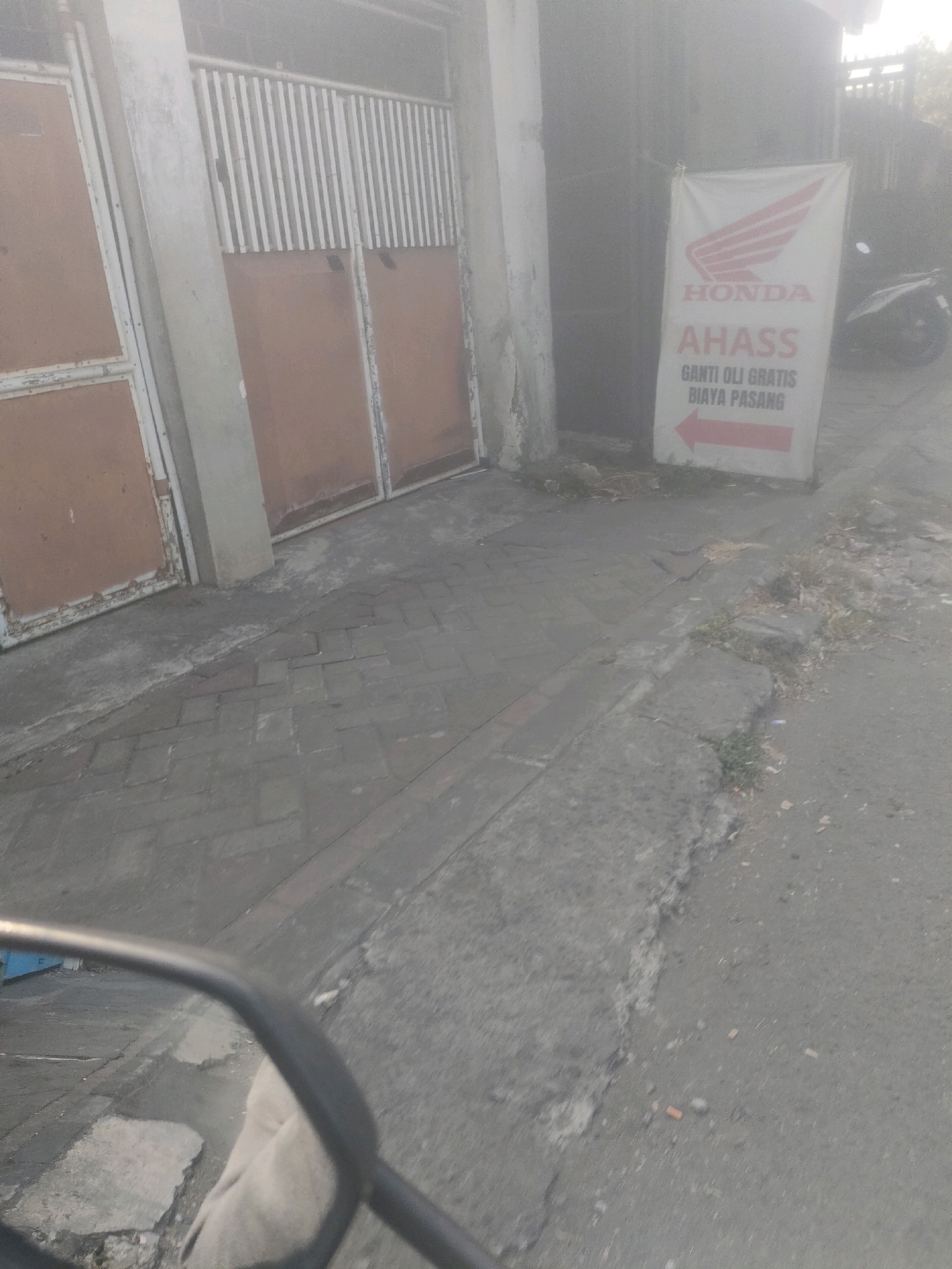 The image depicts a street scene in Surabaya, Indonesia, characterized by a slightly hazy atmosphere and muted lighting, suggesting it may be early morning or late afternoon. In the foreground, a motorcycle's rearview mirror is visible on the left, reflecting a portion of the street and pavement. The pavement itself is a mix of concrete and brick paving stones.
Dominating the middle ground is a beige Honda AHASS banner, which reads "AHASS GANTI OLI GRATIS BIAYA PASANG" (AHASS Free Oil Change, Installation Fee Included), indicating a motorcycle service center. The Honda logo is prominently displayed at the top of the banner, with a red arrow pointing left. To the left of the banner, a large, weathered metal gate with brown panels and a white-barred upper section is visible. The gate appears to be part of a building or property.
In the background, a motorcycle is partially visible to the right of the banner. The environment suggests an urban or semi-urban setting, with a sidewalk and a road. Debris and vegetation are scattered along the edge of the sidewalk, adding to the sense of an everyday street environment. The overall impression is that of a typical commercial street in a Southeast Asian city.