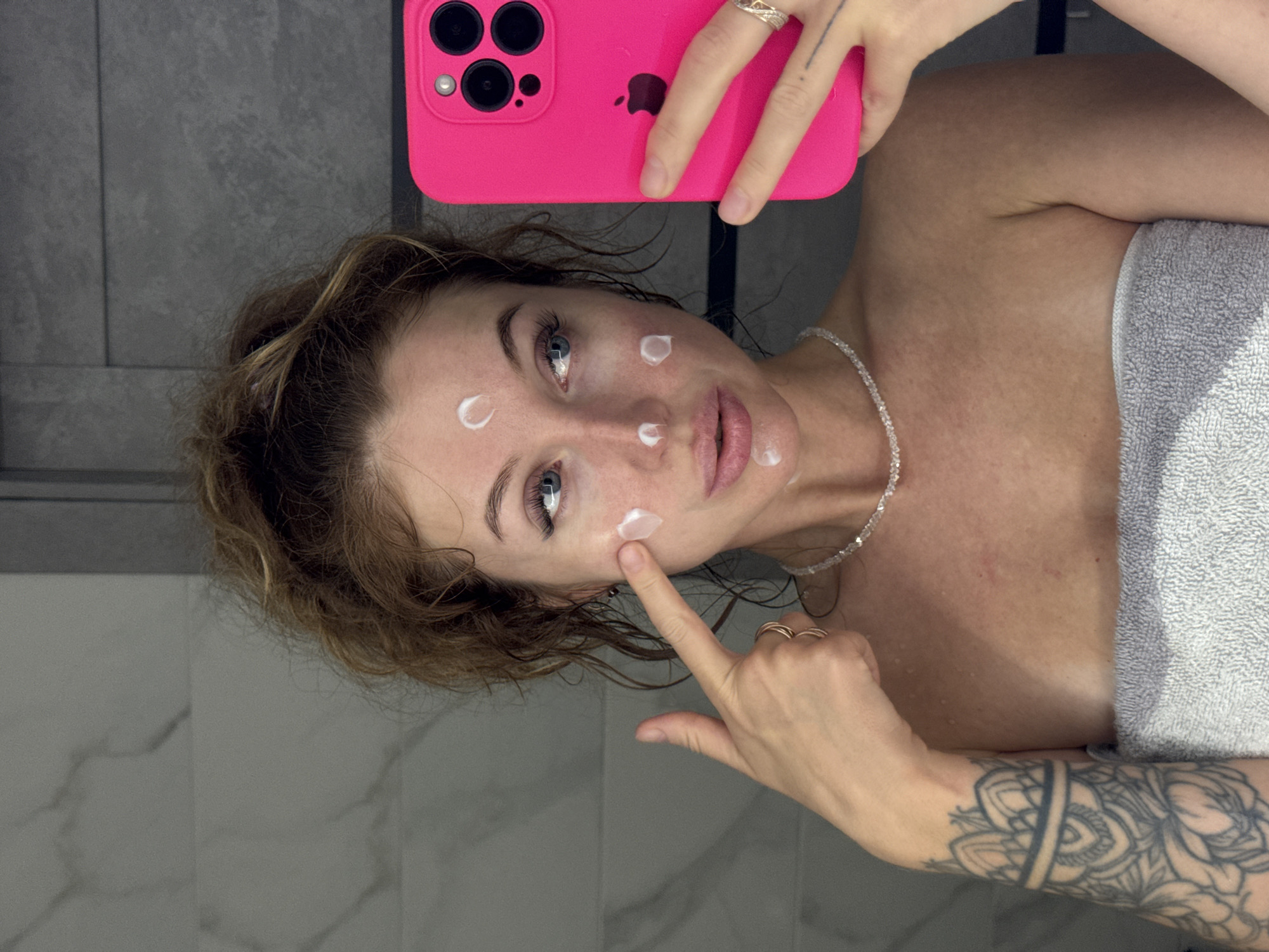 The photograph shows a woman in a bathroom. The background is a grey tiled wall. She is holding a pink smartphone up to take a selfie. She is wearing a grey towel and a beaded necklace. She has several dots of white cream on her face. Her right hand is touching her face and her left hand holds the phone. She has tattoos on her right arm. The image does not provide information about the location in the city of Chaikovsky, Russia.