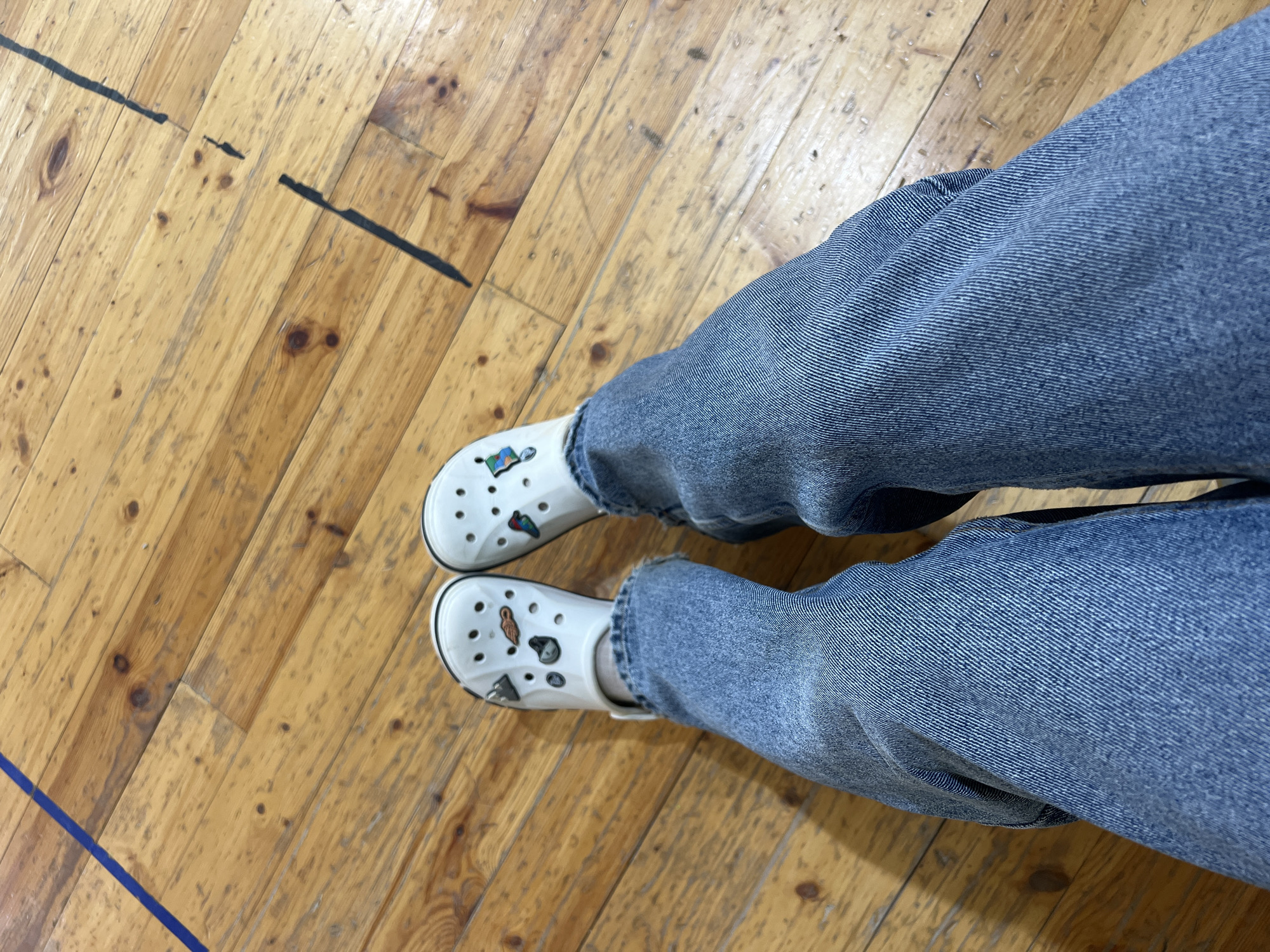 The image captures a top-down view of a person's lower legs and feet, clad in blue denim jeans and white Crocs. The Crocs are adorned with multiple colorful Jibbitz charms, featuring various small designs, though none are distinctly identifiable as text. The person appears to be either standing or sitting on a well-worn, light-colored wooden floor, characteristic of an indoor athletic facility, gymnasium, or studio. The floorboards show natural wood grain, knots, and some signs of wear and tear, including scuffs and dark marks. Distinct black and blue lines, typical of court markings, are visible on the floor, reinforcing the impression of a sports or communal indoor space. The lighting suggests an indoor environment, but the time of day and weather are not discernible. The scene is located in Chaikovsky, Russia. No specific activity beyond being present in the space is depicted, and no emotions can be inferred from the visible parts of the subject.