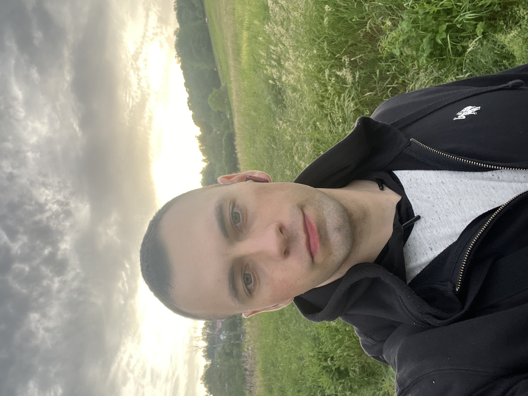 A man is in a field of grass under a cloudy sky, wearing a black hoodie with a white logo and a white shirt underneath. He is looking at the camera. The location is Unknown, Unknown.