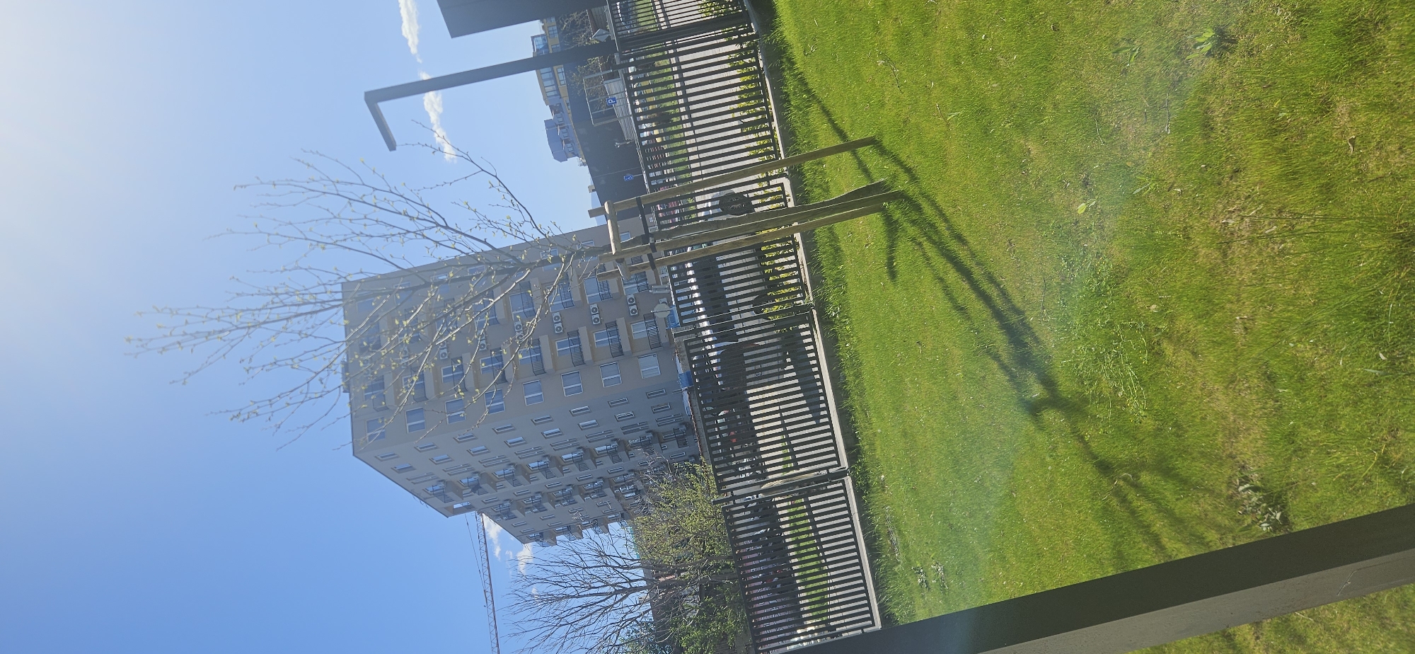 The image shows an exterior scene in Plovdiv, Bulgaria. The foreground is dominated by a green lawn with some shadows cast upon it. A metal fence runs parallel to the lawn. Behind the fence is a street with parked cars and a modern apartment building. The building is light grey and has numerous windows and air conditioning units visible on its facade. A tree with bare branches is partially obscuring the building. The sky is blue with a few white clouds. A streetlight pole is visible in the upper left corner. The image appears to have been taken from an elevated position, possibly from a balcony or upper floor of a building.