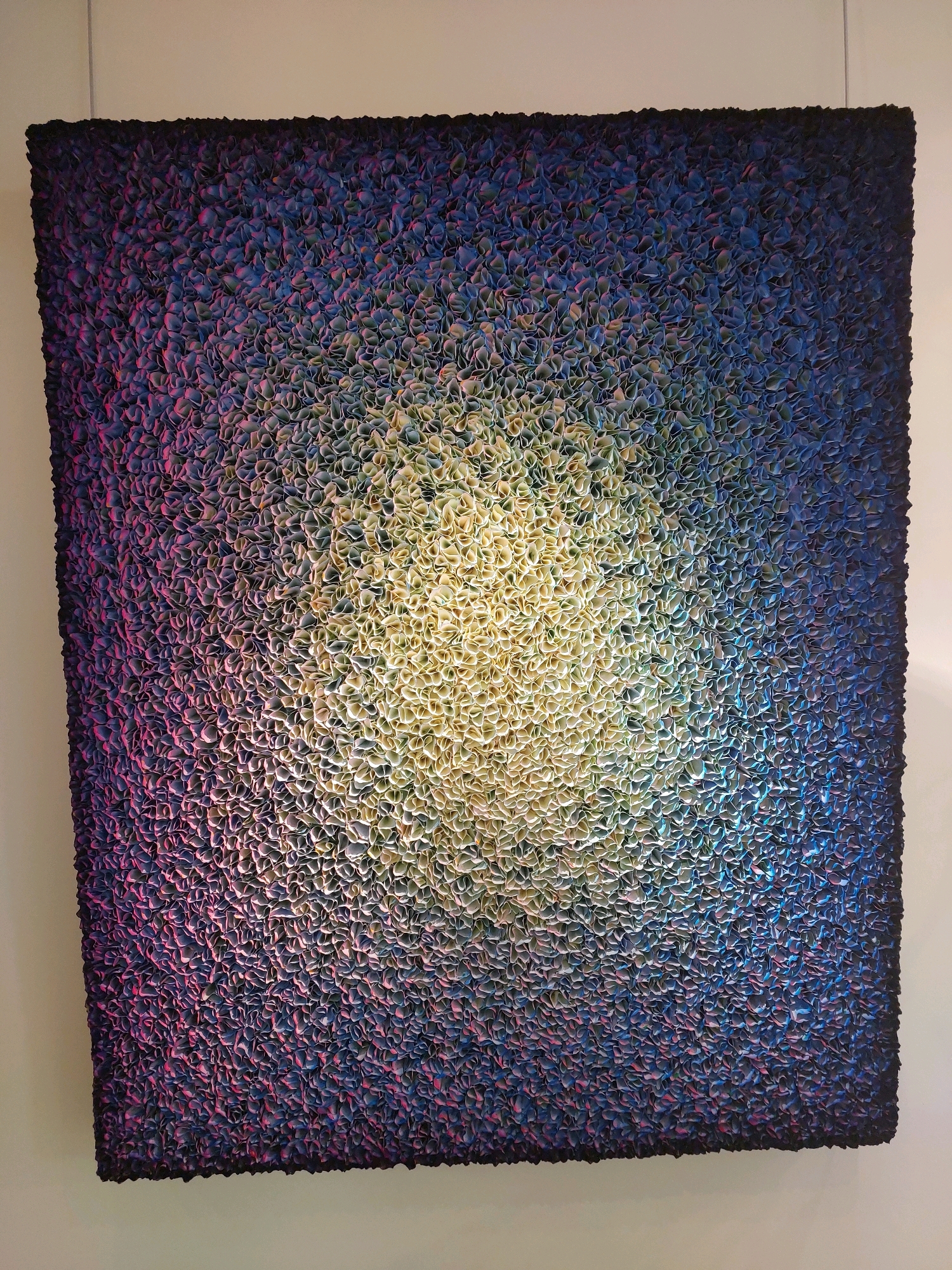 This is a detailed description of an artwork titled "Cosmic Bloom" by French artist Daniel Buren, located in the Maeght Foundation in Saint-Paul-de-Vence, France.
The artwork is a large, textured painting with a gradient of colors. The center of the painting is a bright, almost white-yellow, which gradually fades outwards into shades of light yellow, green, blue, and finally deep purple and black at the edges. The surface of the painting is made up of thousands of small, folded pieces of paper or fabric, giving it a highly textured, almost three-dimensional appearance. These elements are arranged in a dense, organic pattern that resembles a blooming flower or a celestial phenomenon. The light source appears to be coming from the center of the artwork, illuminating the lighter colors and casting subtle shadows on the textured surface. The artwork is hung against a plain, off-white wall, with a thin wire visible at the top suggesting it is suspended. There are no people or other objects visible in the immediate vicinity of the artwork. The Maeght Foundation is known for its impressive collection of modern and contemporary art, set within beautiful gardens, offering a serene environment for viewing art.