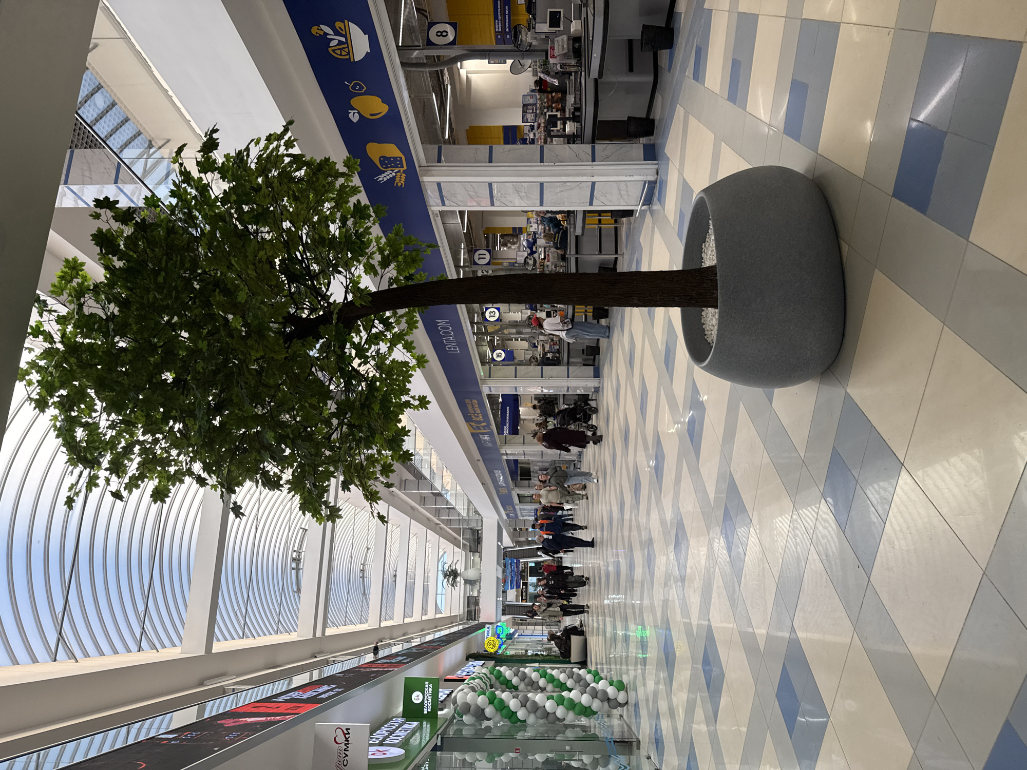 The image shows an indoor scene at a Lenta hypermarket in Izhevsk, Russia. The store features a large faux tree planted in a gray, pot-like structure. The floor is tiled with a diamond pattern in light blue and white. A row of checkout lanes is visible in the background, with customers and employees present. Banners and signage are displayed above the lanes. The ceiling has a curved, architectural design with glass panels. A display of green and white balloons is located near the bottom of the image. People are walking along the aisle in the middle of the store.