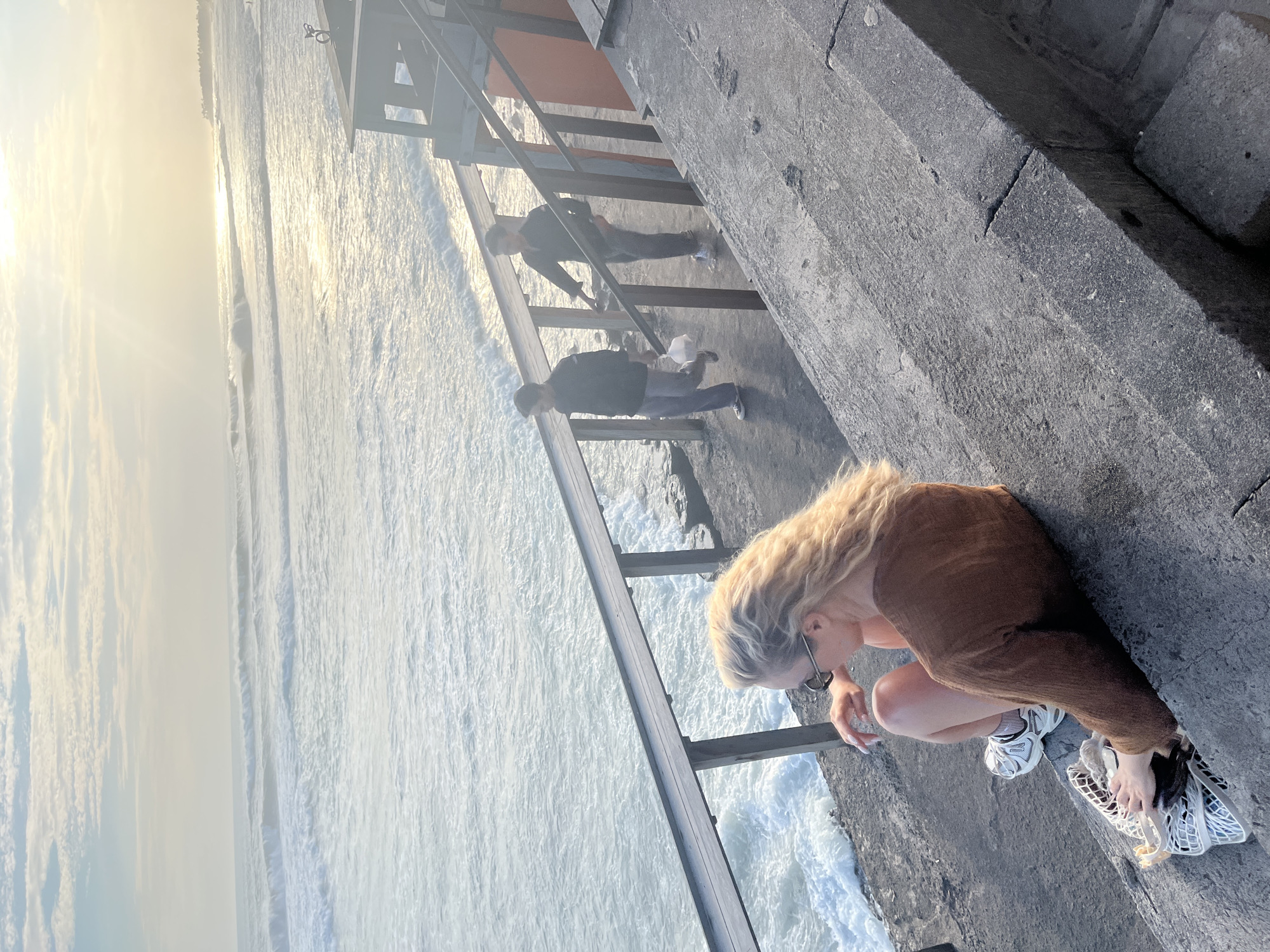 A woman with blonde hair is sitting on concrete steps overlooking a choppy ocean, with waves breaking near the shore. She is wearing a brown sweater, shorts, and white sneakers. Next to her is a white net bag containing items. Two men are walking up a wooden walkway with a metal railing to the right of the woman. The sun is low in the sky, casting a warm glow over the scene. The location appears to be a coastal area in North Kuta, Indonesia.