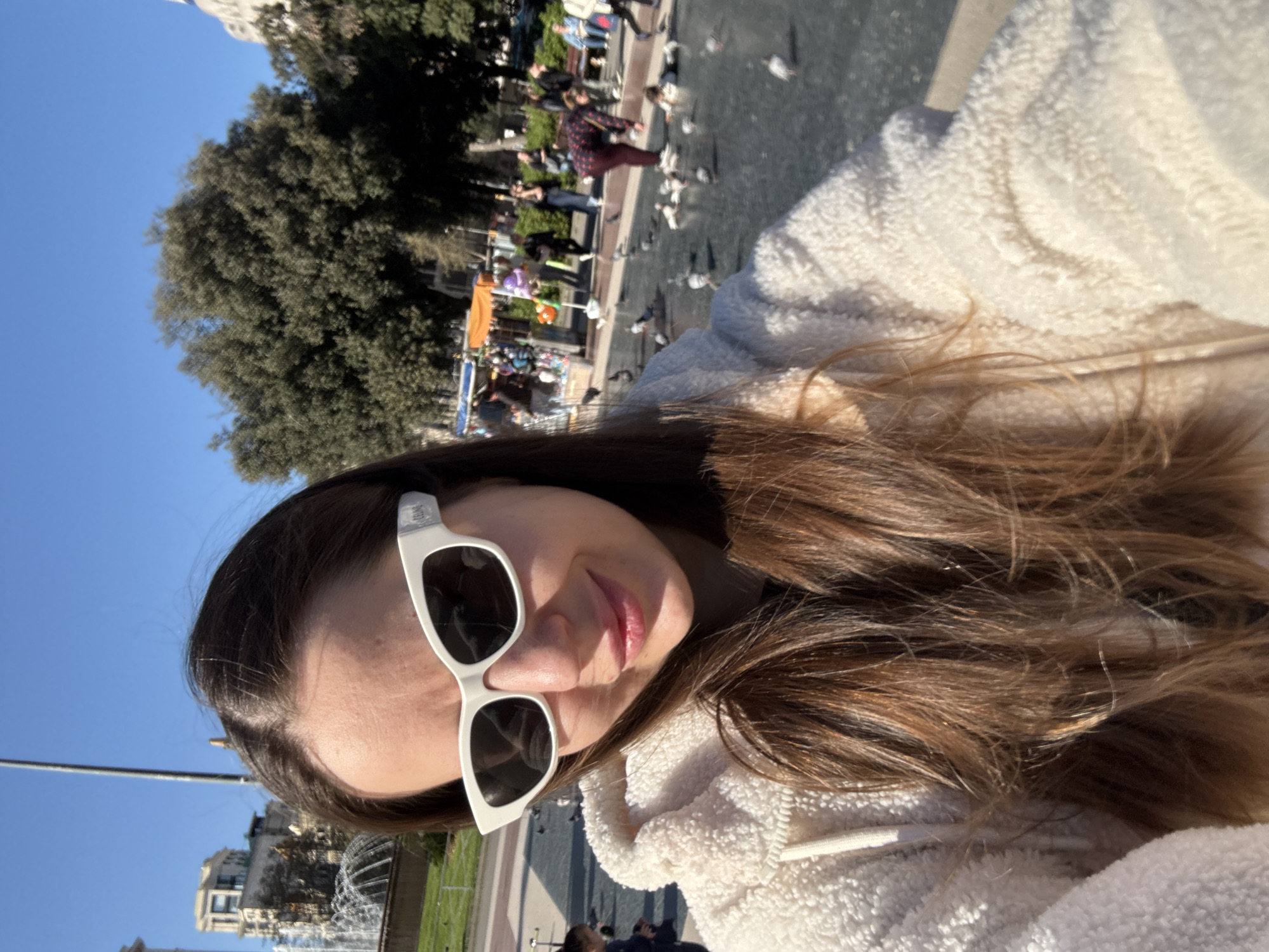A woman wearing white sunglasses and a fuzzy white jacket is posing for a selfie in a sunny outdoor location. In the background, there is a cityscape that includes buildings, a fountain with water spraying upwards, trees, and a clear blue sky. Further in the background, people are walking and interacting with pigeons on a paved area. The location appears to be in a public square or park setting. Considering the architecture and general atmosphere, this scene is likely in Barcelona, Spain.