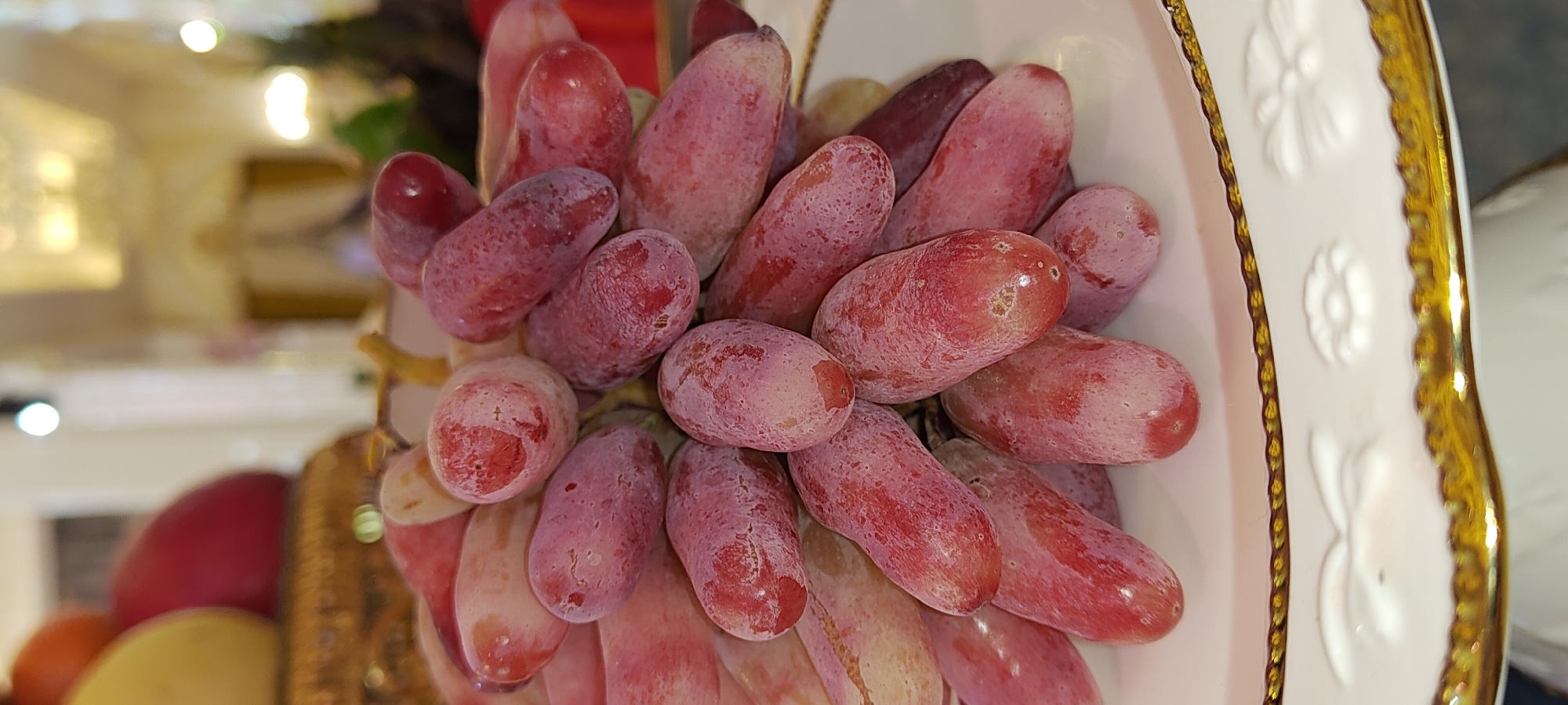 A bunch of red, elongated grapes with a dusty coating is presented on a white plate with a gold trim. The grapes are arranged in a cluster, filling most of the frame. The plate has embossed floral patterns along its rim. In the blurred background, there are hints of other fruits, possibly mangoes, and a dimly lit indoor setting with decorative elements, suggesting an event or a display. The scene appears to be a still life composition. There are no people or animals visible in the image. The specific location in Khujand, Tajikistan cannot be determined from the visual information provided.