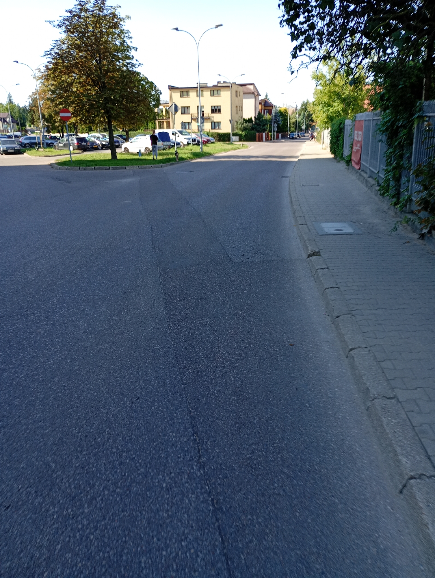 The image depicts a street scene in Białystok, Poland. In the foreground, a dark asphalt road curves to the right. To the left of the road, a circular grassy area is visible with a mature tree and parked cars. A red and white circular stop sign is placed on the grassy area. Further in the background, a light yellow building with multiple windows stands along the street. To the right of the road, a paved sidewalk runs alongside a gray metal fence, with green foliage growing on the fence and above it. A person in dark clothing is seen walking on the sidewalk in the mid-ground. The sky is clear and blue, indicating daytime.