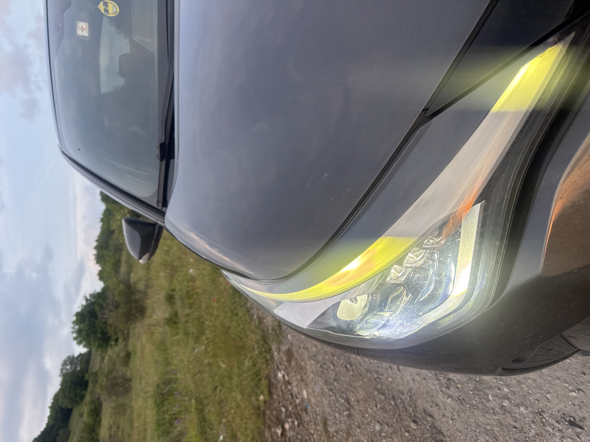 The image depicts the front-right quarter of a dark grey vehicle parked on an unpaved dirt track in a rural or semi-rural setting in Dragovishtitsa, Bulgaria, under an overcast sky.
The vehicle's front-right headlight is illuminated, featuring a white light from the main projector lens and a prominent, curving amber/yellow LED strip as a daytime running light. Dust and dirt particles are visible on the headlight housing and the adjacent bodywork. The dark grey hood surface reflects the dull sky and shows smudges or dust. The upper portion of the front-right tire and wheel well, heavily soiled with dirt, is partially visible at the bottom left.
Through the reflective windshield, a yellow smiley face charm with sunglasses hangs from the interior rearview mirror area, and a red cross sticker (first aid symbol) is affixed to the upper right corner. The black windshield wiper blade rests at the base of the windshield. The black right side mirror is partially visible on the far right.
The immediate ground is an unpaved dirt track composed of light brown soil and small stones. Along the right edge of the track, sparse green grass and some small, low-lying green plants, including hints of purple flowers, grow.
In the midground and background, gentle green hills rise, densely covered with a mix of trees and shrubs, indicating a vegetated landscape. The sky above is overcast with light grey clouds, suggesting diffused natural light without direct sunlight. No people or animals are visible in the scene.
