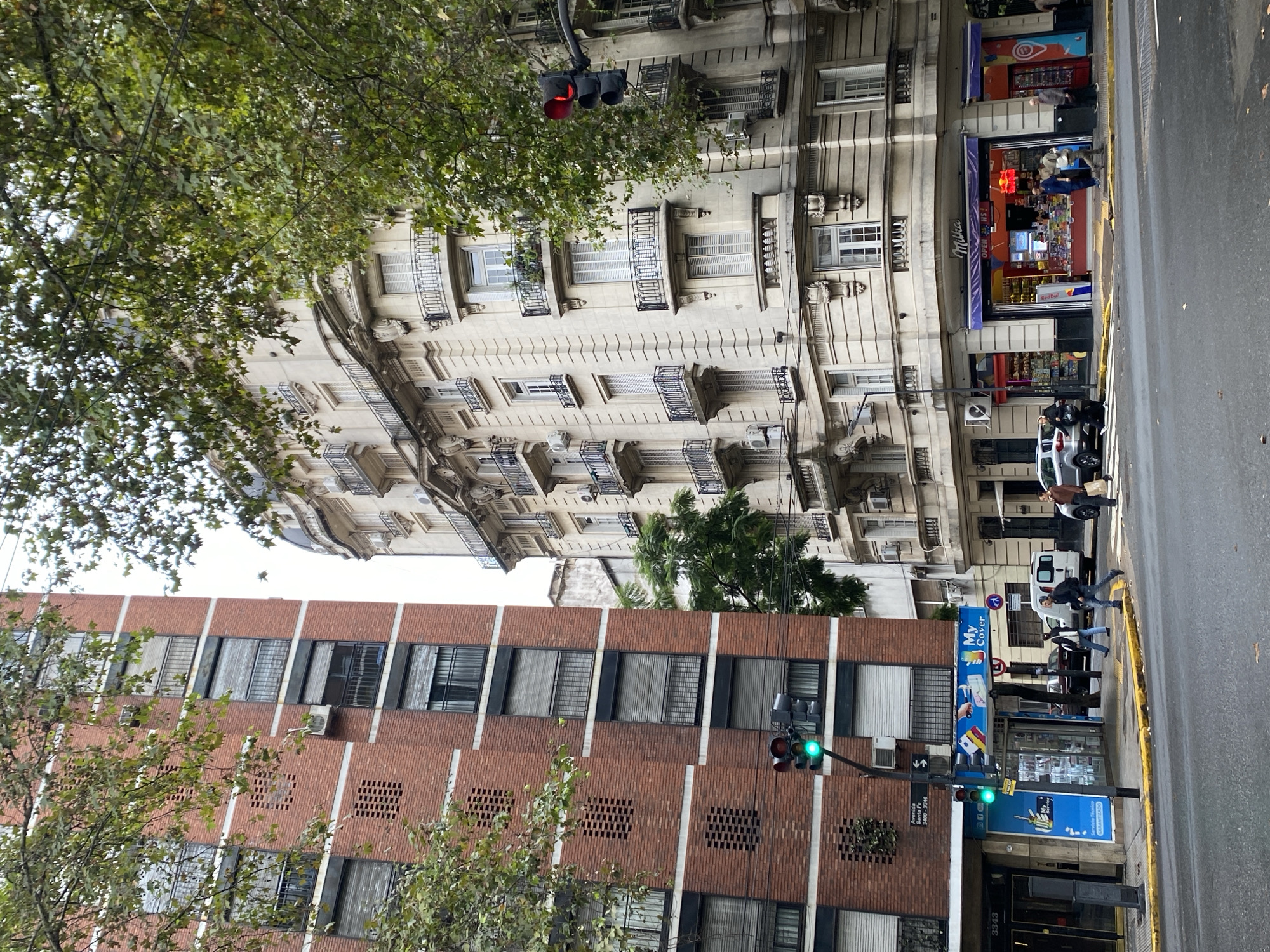 The image depicts a street scene in Buenos Aires, Argentina. On the right side of the frame, there is a multi-story building with a light-colored facade, featuring balconies and windows. Beneath it, a small store is visible, and a person is standing in front of it. People are walking across the street. Cars are parked along the curb. On the left side, there is a brick building with windows. A street sign indicates the location is Avenida Santa Fe 3400-3340. Traffic lights are visible, with one showing a green light and the other a red light. Trees with green leaves frame the upper portion of the image.