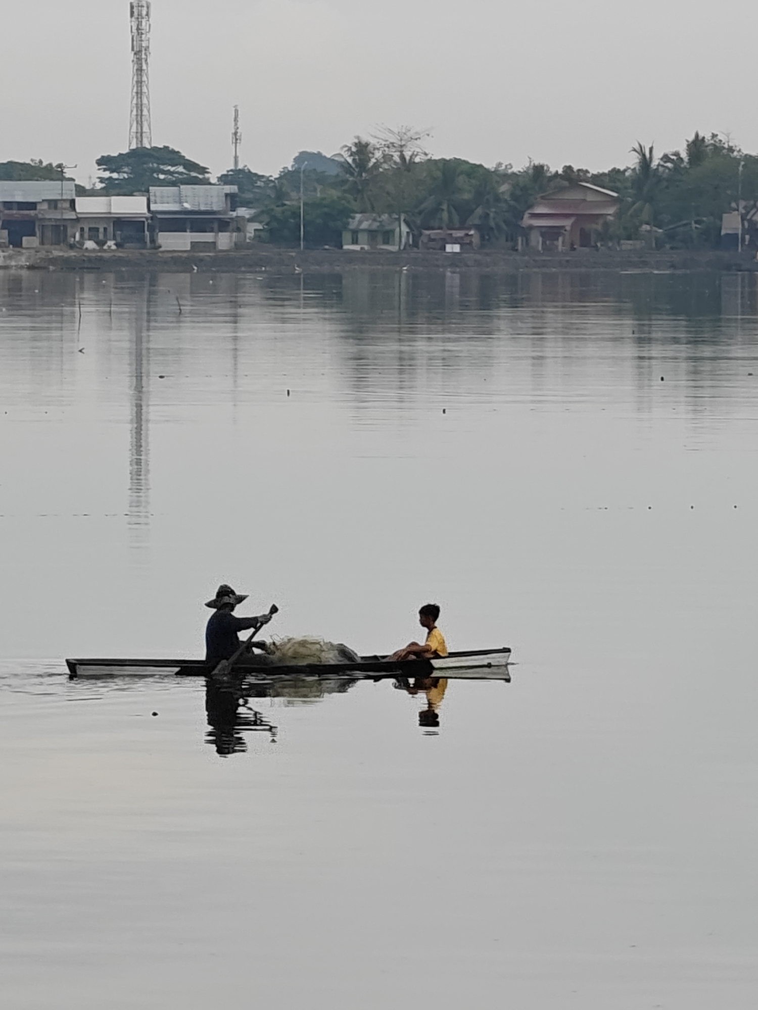 An overcast day shows a calm body of water reflecting the sky, houses, and trees on the shore. A narrow, wooden boat with two occupants is in the foreground. The person in the rear, wearing a wide-brimmed hat and dark clothing, is rowing with a paddle. A net is piled in the boat behind them. In the front of the boat, a young person in a yellow shirt sits facing forward. The shoreline in the background features a cluster of houses and buildings, with two prominent communication towers rising above the trees. The overall scene suggests a peaceful, everyday moment in a waterside community, likely in Lhokseumawe, Indonesia, given the presence of communication towers and the style of buildings.