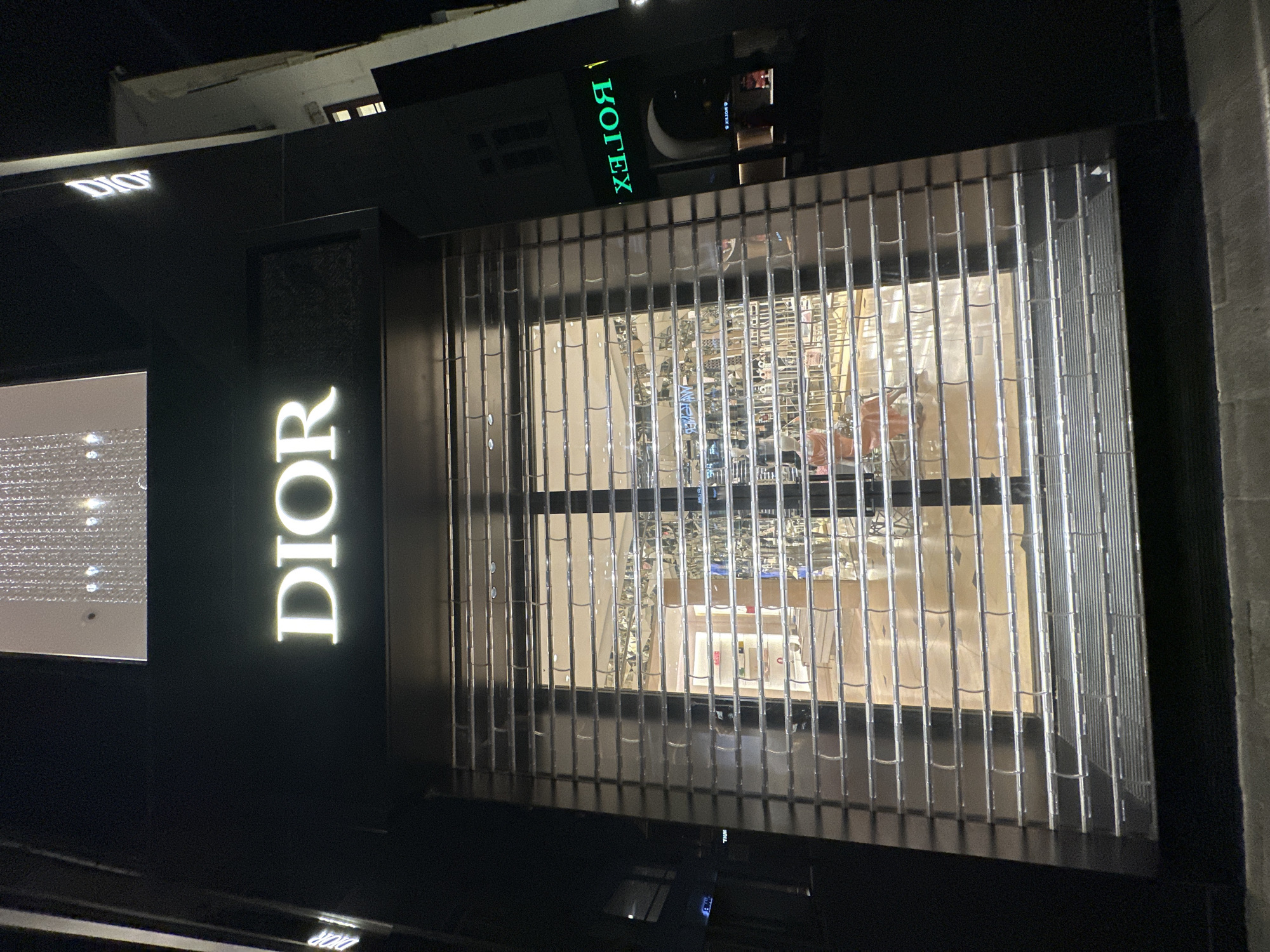 The image shows the exterior of a closed Dior store in Vienna, Austria. The store is illuminated from within, and the large storefront window is covered by a metal security grate. Above the window, the Dior logo is prominently displayed in large, glowing white letters. To the right of the Dior sign, another sign reads "ROLEX" in green illuminated letters. The surrounding environment is dark, suggesting it is nighttime. The ground in front of the store is paved.