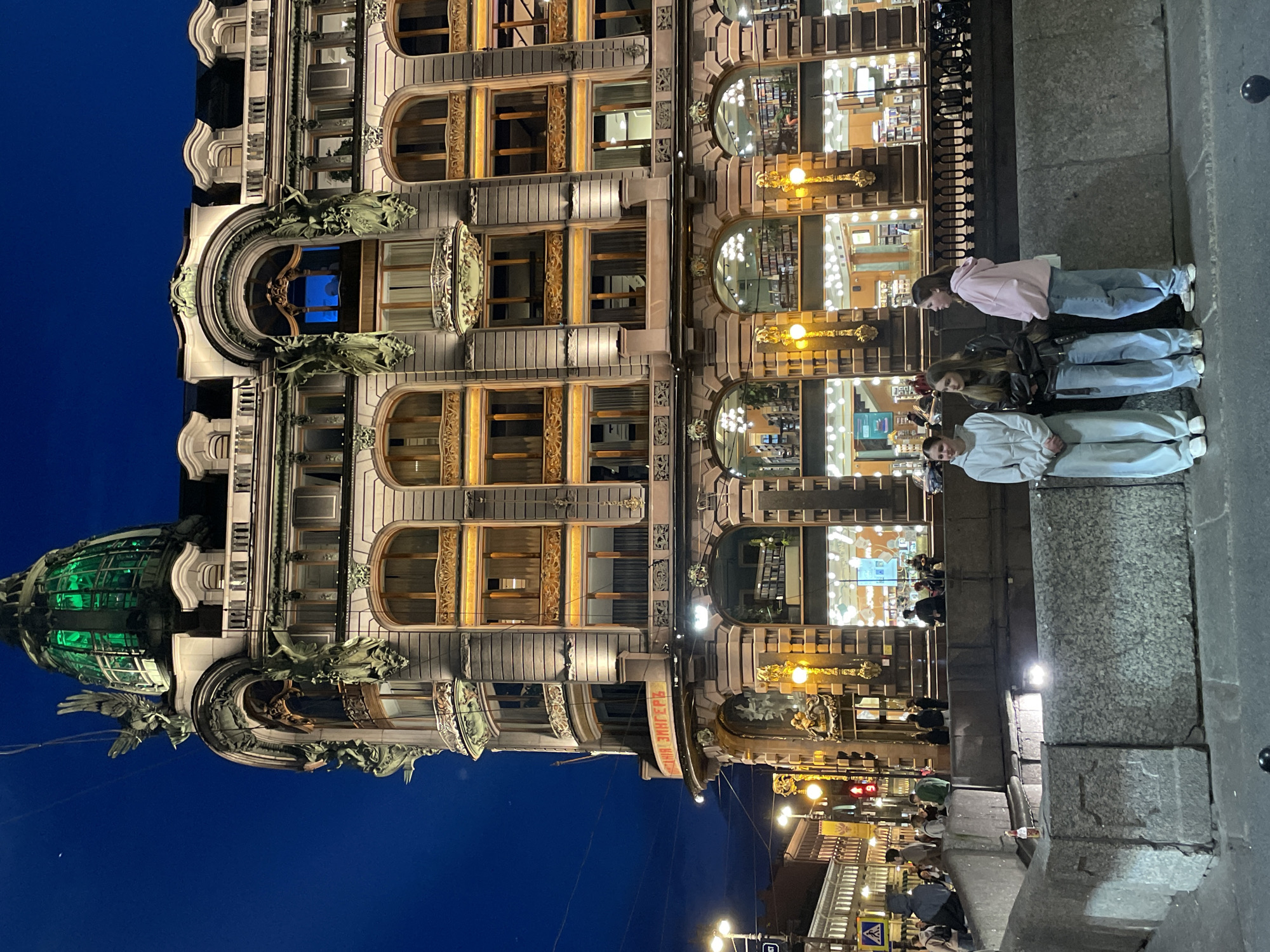 The image displays a grand, ornate building in an urban setting at night, strongly identifiable as the Singer House (Dom Knigi) in Saint Petersburg, Russia. The sky above is a dark, clear blue, indicating late evening or night.
The multi-story building, an exemplary piece of Art Nouveau architecture, dominates the left side of the frame. Its facade is richly decorated with intricate details, including large arched windows, some featuring elaborate golden-colored frames and balconies. Numerous sculptures, resembling gargoyles and winged figures, adorn the building's cornices and ledges. A distinctive, large glass dome, illuminated with a vibrant green light, crowns the building's upper left section. Many of the building's windows glow brightly from within, revealing retail interiors, consistent with the Cyrillic text "ДОМ КНИГИ ЗИНГЕРЪ" (Dom Knigi Singer, meaning Singer House of Books) visible on a curved sign or balcony midway up the facade.
On the right, a wide stone embankment or sidewalk runs parallel to the building. A group of three young women stands on this pedestrian area, facing the illuminated building. One woman wears a pink hoodie and light blue jeans, another a black leather jacket with light blue jeans, and the third a light grey hoodie and sweatpants. They appear to be observing the building or posing. Other indistinct figures are visible further down the street and inside the brightly lit building, suggesting an active urban environment. Streetlights cast warm glows, further illuminating the scene. The overall atmosphere is calm and bustling, characteristic of an evening in a vibrant city center.
