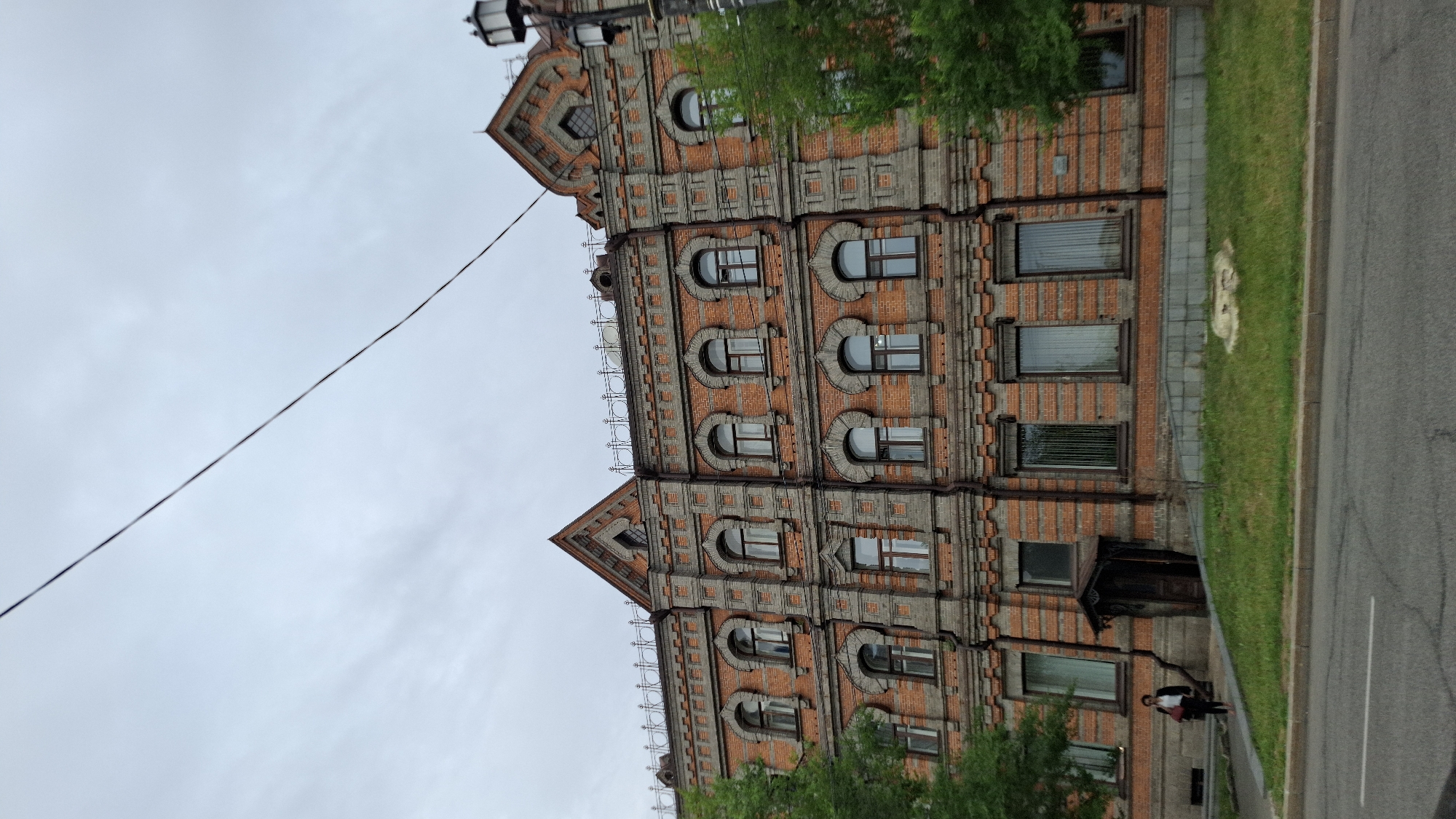 The image captures a facade of a red brick building with ornate grey stone accents and arched windows, typical of early 20th-century architecture. The building is situated on a street in Khabarovsk, Russia, as indicated by the presence of a woman walking on the sidewalk in front of it. The woman, dressed in dark clothing and carrying a red bag, is positioned to the left of the building's entrance. The sky is overcast, suggesting a cloudy day. Trees are visible on the left and right sides of the frame, partially obscuring the building. A street with visible lane markings runs in the foreground. The overall scene depicts an urban environment with a historical building.