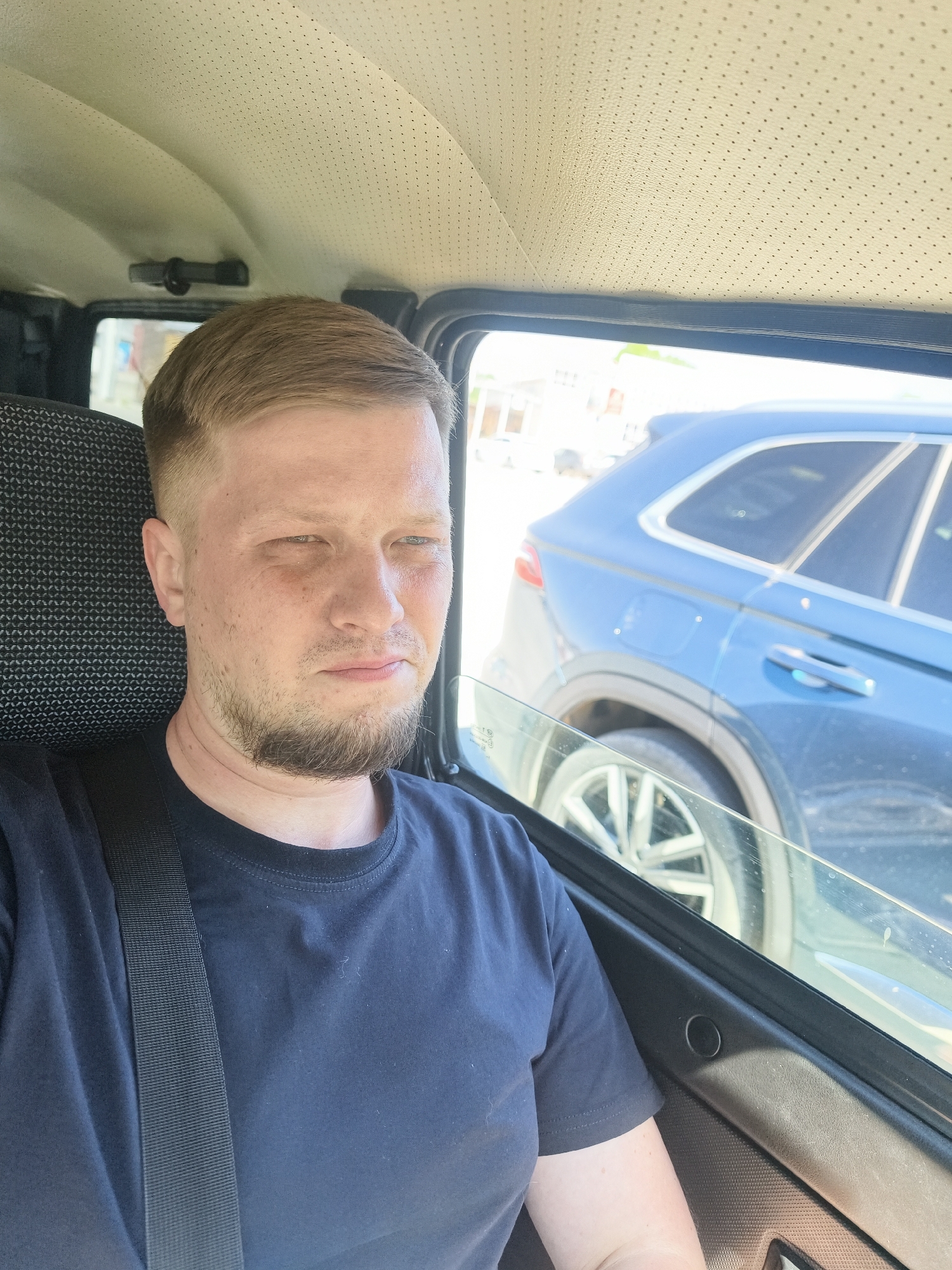 A man sits in the driver's seat of a car, wearing a black t-shirt and a seatbelt. He has short brown hair and a beard. The car is parked next to a blue SUV, visible through the open window. The car's interior has a patterned seat and a perforated headliner. The location is likely Chaikovsky, Russia.