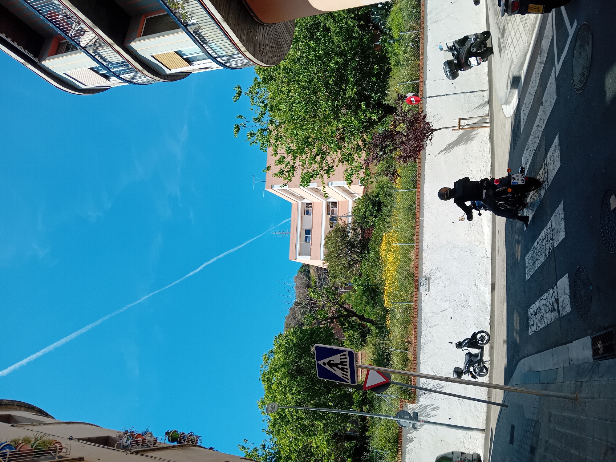 The image captures a vibrant daytime street scene in what appears to be a residential area, rotated ninety degrees clockwise. The sky is a clear, brilliant blue with a single prominent white contrail stretching across it. Sunlight casts strong shadows, indicating a bright, sunny day.
In the foreground, a person wearing a helmet and dark clothing is riding a black motorcycle, moving right to left across a pedestrian crossing. The motorcycle has a visible license plate: "HG 01 XG 74". Another black scooter is parked on the sidewalk to the left of the pedestrian crossing, and a third scooter is parked further down the street to the right. The road features white pedestrian crossing markings and manhole covers.
To the left of the road, a wide, light-colored sidewalk runs alongside a low wall and a metal fence. Beyond the fence is a strip of greenery with lush trees and yellow wildflowers. A blue pedestrian crossing sign and a red-and-white yield sign are visible on the sidewalk. A small white sign attached to the low wall reads "C/ SANT... TARR".
On the far left, partially visible, are sections of residential buildings with balconies and railings. Across the street, in the background, another multi-story residential building with light-colored walls and numerous windows is visible, partially obscured by green trees. Beyond this building, a green, hilly landscape rises, suggesting proximity to natural terrain.