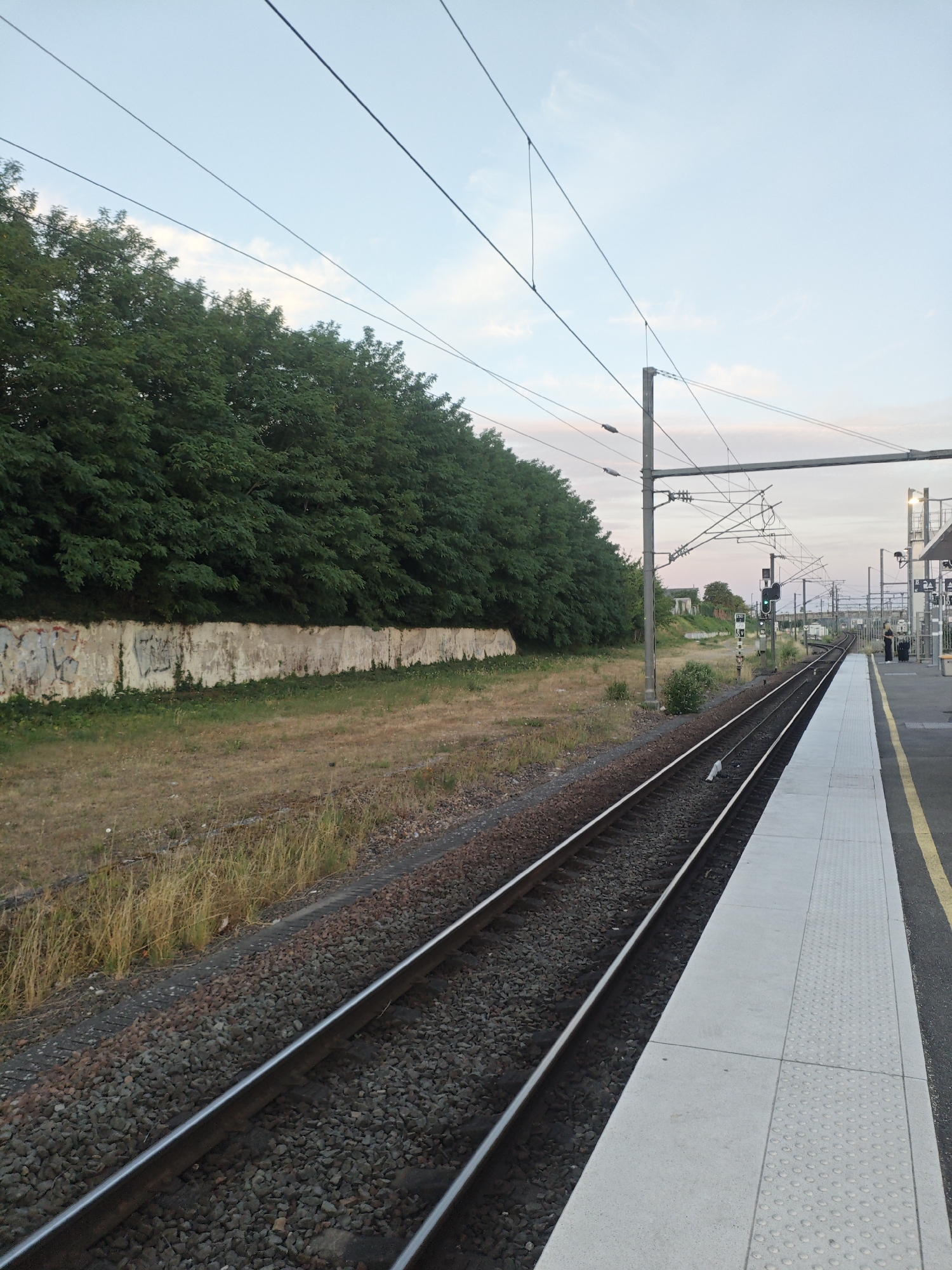 The image depicts a train station platform in Vaires-sur-Marne, France. In the foreground, train tracks run diagonally from the lower left to the upper right of the frame. The platform, on the right side of the tracks, is made of light-colored concrete with a textured safety strip along the edge. A yellow line marks the boundary of the platform. Beyond the tracks, a low, weathered concrete wall is visible, covered in graffiti. A dense line of mature green trees forms a backdrop to this wall. Overhead, numerous electrical wires, part of the catenary system for the train, stretch across the sky, supported by grey poles. The sky is a pale blue with hints of pink and white clouds, suggesting it might be early morning or late evening. In the distance, on the right side of the tracks, a green train signal is illuminated, and two figures can be seen standing on the platform. The overall scene conveys a sense of a functional, somewhat rural, train station environment.