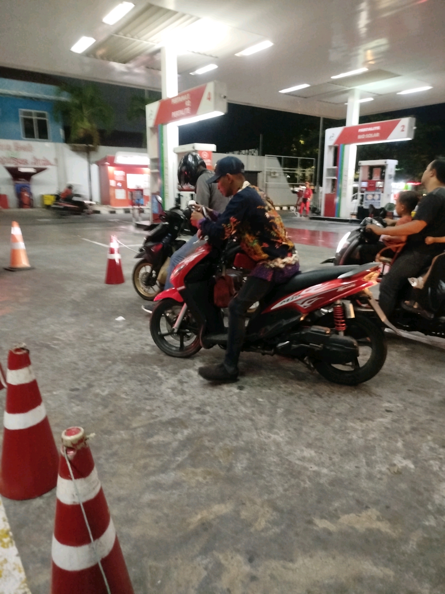 At a brightly lit gas station in Surabaya, Indonesia, a man on a red scooter is captured in the foreground, leaning slightly forward. He wears a patterned shirt and dark pants, with a blue cap on his head. To his right, another scooter is visible, with a person in the background wearing a dark t-shirt and jeans. In the distance, a child is seated on the back of the scooter, facing away from the camera. The gas station's overhead canopy is illuminated by fluorescent lights, and several red and white traffic cones are strategically placed on the wet asphalt ground, indicating a recent rainfall. The background shows elements of the gas station's facilities, including pumps and signage.