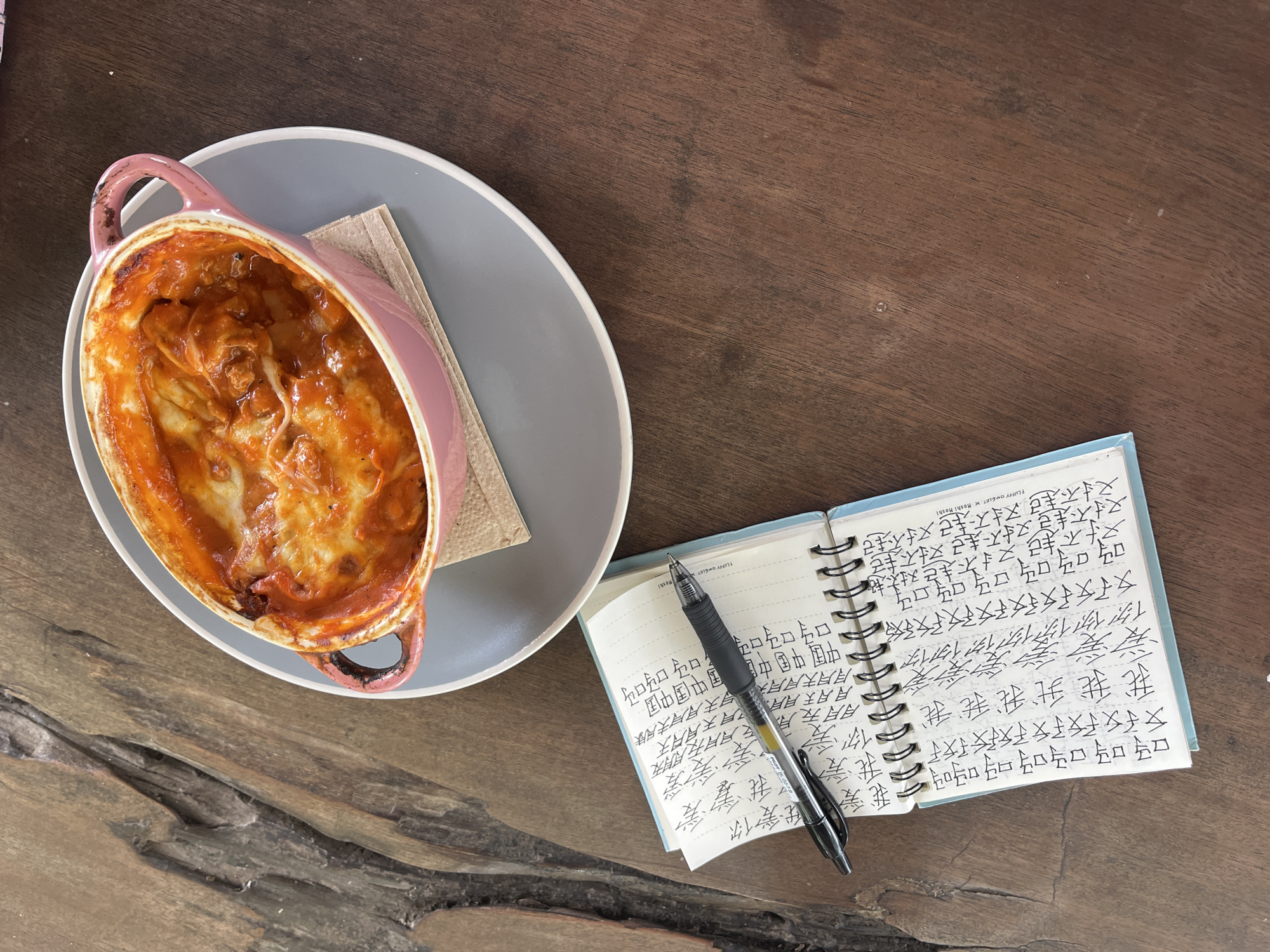 The image shows a tabletop scene on a wooden surface with a gray dish holding a pink baking dish filled with food, likely lasagna or a similar casserole, along with a notebook and pen. The notebook is open and contains writing in a language that appears to be Chinese. The pen rests on the open pages. The image was taken in Ban Tom, Thailand, as it is stated in the provided OCR.