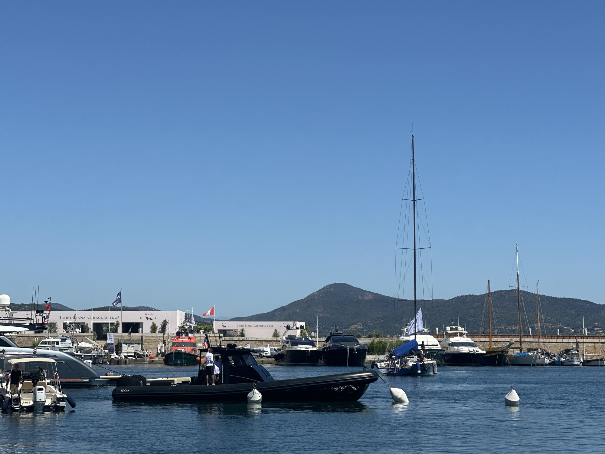 The image depicts a bright, sunny day in a harbor, likely in Saint-Tropez, France, given the context of the Loro Piana Giraglia event. The foreground is dominated by the blue water of the harbor, with several buoys floating on the surface. In the middle ground, a black rigid inflatable boat (RIB) with a partially visible name "TOOKA" is prominently featured, with two individuals on board. To the left of the RIB, a smaller white motorboat, possibly named "JAWS," is moored. Behind these boats, a variety of other vessels are docked, including larger yachts, sailboats with tall masts, and a distinctive orange and green rescue boat.
In the background, a long, light-colored building with the text "LORO PIANA GIRAGLIA 2026" is visible, adorned with several flags, including French and Italian tricolors. Beyond the building, a lush green, tree-covered mountain rises against a clear blue sky. The overall scene conveys a sense of a vibrant maritime event or gathering, characterized by the presence of numerous boats and a festive atmosphere implied by the event signage.