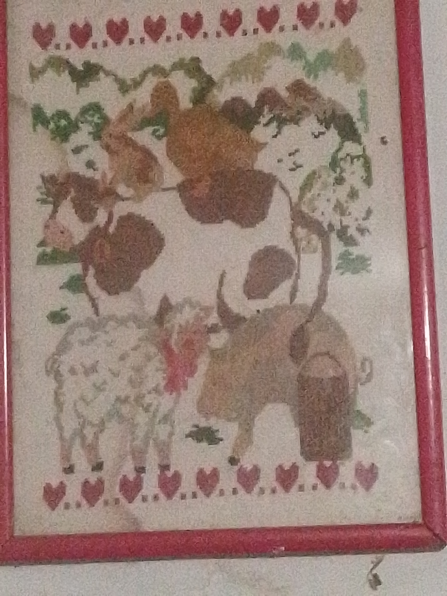 The image is a close-up, slightly blurry photograph of a framed piece of embroidered artwork. The artwork depicts a pastoral scene with several farm animals, including a cow, a sheep, and possibly a bull. The animals are rendered in a stylized, pixelated manner, typical of cross-stitch or needlepoint. Above the animals, there are green leafy elements that could represent trees or bushes, and small white dots suggesting flowers. The top and bottom borders of the artwork are decorated with a repeating pattern of red hearts and small dots. The frame is a simple, solid red color.
The image was taken in Santa Ana, Venezuela.