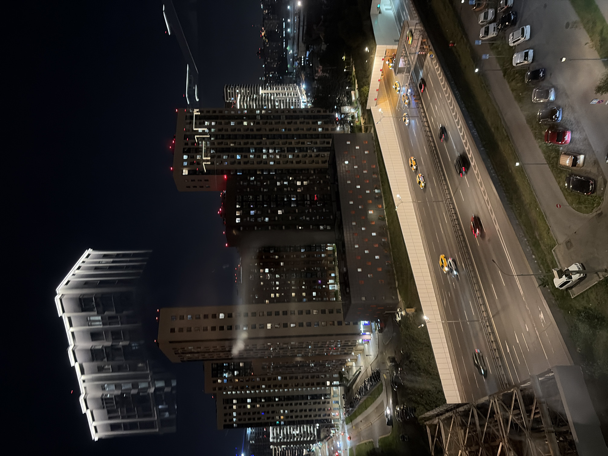 This is a nighttime, high-angle, long-shot photograph of a modern city district in Moscow, Russia, likely taken from a high-rise building. The image captures a complex urban landscape dominated by towering residential buildings, illuminated by numerous lit windows, suggesting a densely populated area. In the foreground, a multi-lane highway is visible, with cars, including yellow taxis, traveling at speed. A tunnel entrance is also present on the highway. The buildings are of contemporary architectural design, featuring clean lines and extensive glass facades. Some buildings have distinctive lighting features on their upper levels. The sky is dark, indicative of nighttime, with no visible stars or moon. The overall scene depicts a bustling urban environment at night, with the infrastructure of a major city clearly visible.