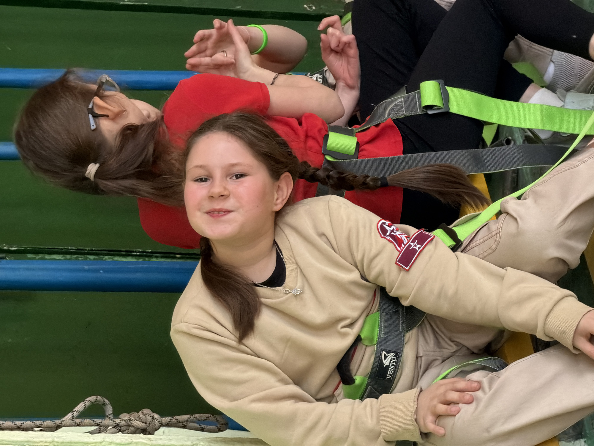 This image captures two females engaged in an adventure activity, likely at a rope course or similar facility in Izhevsk, Russia. The perspective is slightly overhead, showing the subjects leaning or suspended, secured by safety harnesses.
In the foreground, a young girl with a long, brown braid smiles directly at the camera, exuding happiness and confidence. She wears a light beige sweatshirt, featuring a red and white patch on her right sleeve, and matching beige pants. A delicate silver necklace with a small pendant is visible around her neck. She is fitted with a gray harness accented with neon green straps and buckles, clearly displaying the brand name "VENTO" and its logo on a black patch.
Behind and partially obscured, an older female, possibly an adult, is visible. She has brown hair pulled back, wears glasses, a red t-shirt, and black pants. Her left wrist sports a neon green bracelet. She is also secured in a similar gray and neon green safety harness. Her expression is not fully visible, but she appears relaxed in her position.
The background features a solid green wall or surface, traversed by a prominent blue horizontal rail or bar. In the lower left corner, a thick, braided grey rope with a visible knot reinforces the nature of the activity. The bright expressions, particularly from the foreground subject, suggest an enjoyable and engaging experience within this adventure setting.