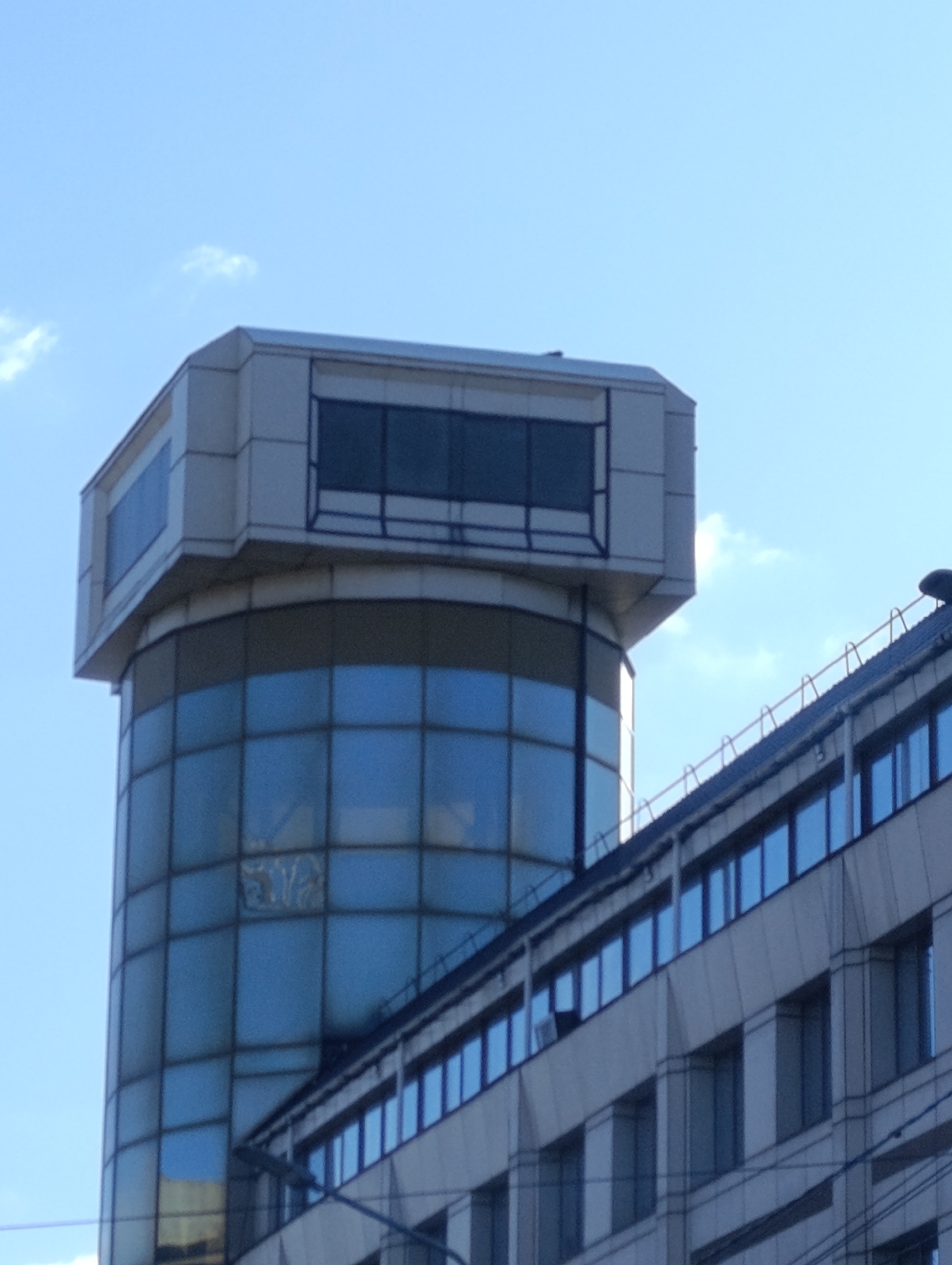 The image shows a tall building in Krasnoyarsk, Russia. A cylindrical glass tower with a box-like structure on top rises above the adjacent building. The glass tower is divided into a grid of windows, reflecting the blue sky. The adjacent building is lower, with horizontal rows of windows. The sky is clear with a few scattered clouds.