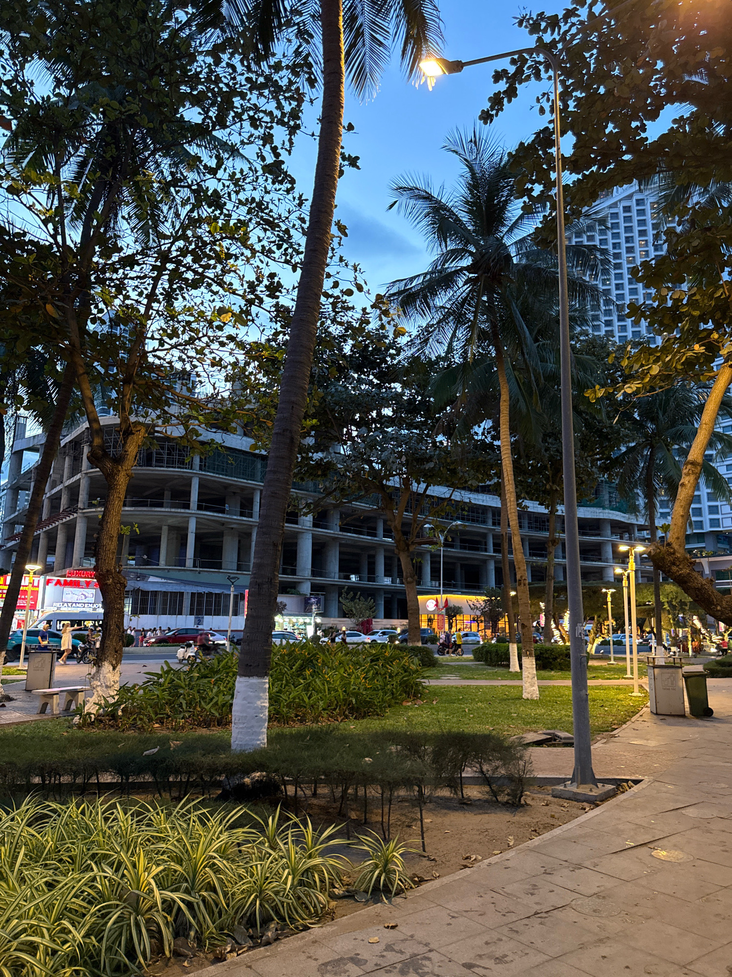 The image shows a street scene in Nha Trang, Vietnam, likely at dusk or dawn, given the twilight sky and illuminated streetlights. In the foreground, a paved walkway runs alongside a grassy area with manicured hedges and ornamental plants. Palm trees and other tropical foliage are prominent throughout the scene.
In the background, a large, unfinished concrete building under construction dominates the cityscape. Its multi-story structure reveals exposed concrete floors and columns, with scaffolding visible in some sections. Across the street, there is a line of businesses with illuminated signage, including one that reads "LUXURY." A variety of vehicles, including cars, motorcycles, and a white van, are parked or in motion on the road. Several people are visible, some walking on the sidewalk, while others are near the vehicles.
Streetlights, including a tall pole-mounted one with a visible bulb, illuminate the area. Trash receptacles are positioned along the walkway. The overall impression is of a developing urban environment with a blend of natural tropical elements and ongoing construction.