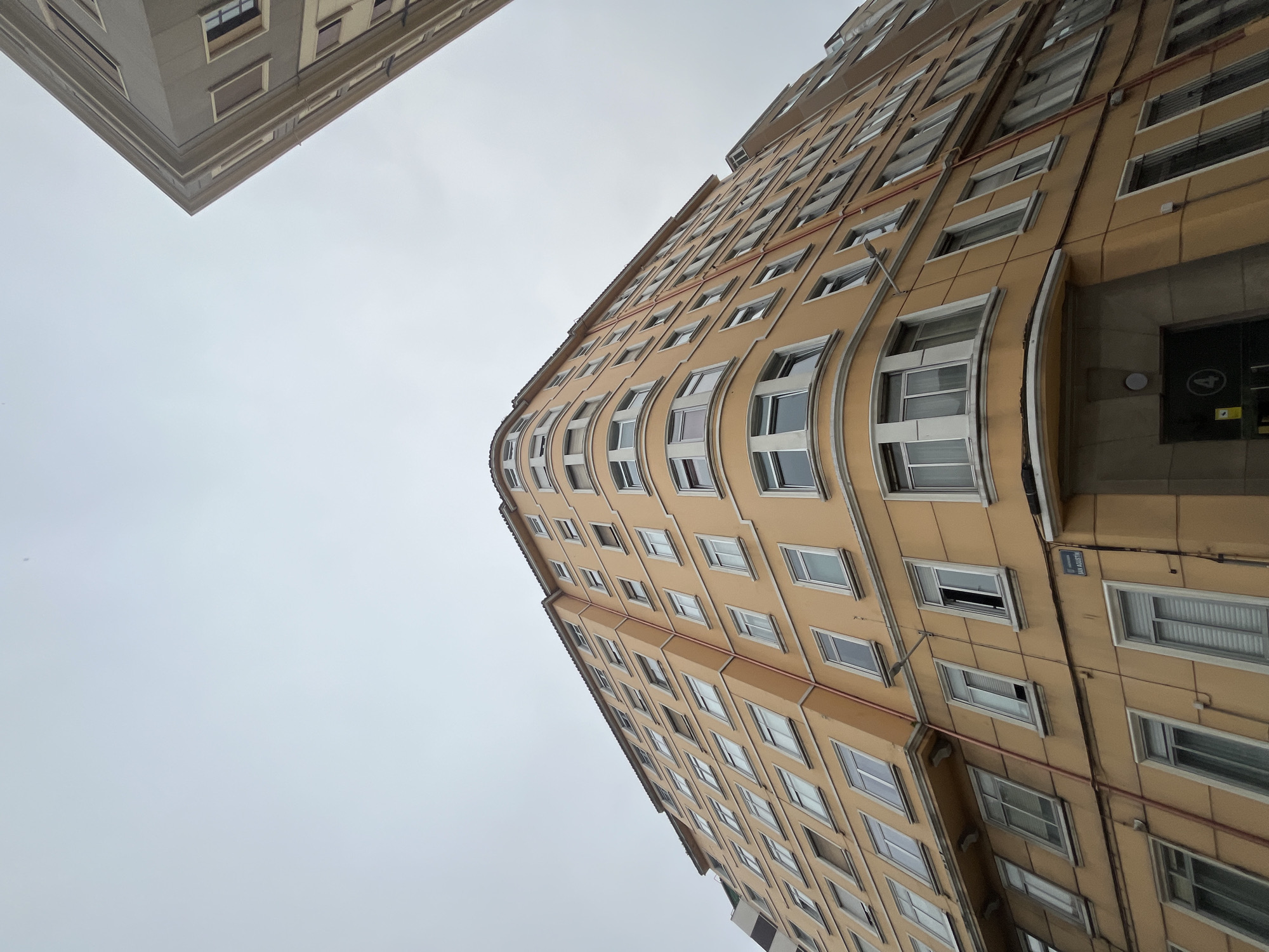 The image shows a tall, light-brown building in A Coruña, Spain, viewed from a low angle. The building has numerous rectangular windows arranged in a grid pattern. The building curves slightly outward in the middle. A second, grey building is visible in the upper-left corner of the frame. The sky is overcast. A pipe or conduit runs vertically along the facade of the light-brown building. A sign is visible on the facade near the bottom right.